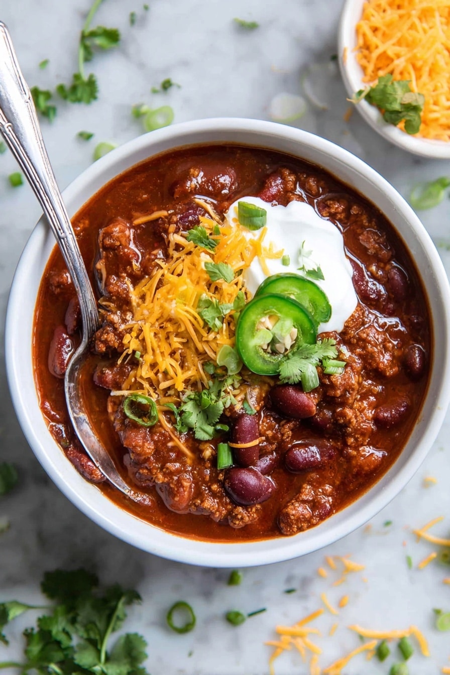 The image shows a white bowl filled with thick chili made of dark red beans and brown meat in a deep red sauce. On top, there is a layer of bright orange shredded cheese, a dollop of white sour cream, and green chopped spring onions with a slice of green jalapeño pepper and fresh cilantro leaves. A silver spoon is placed inside the bowl. The bowl sits on a white marbled surface with some scattered shredded cheese and green herb sprigs nearby. Photo taken with an iphone --ar 2:3 --v 7 - Healthy Ground Turkey Chili, ground turkey chili, healthy chili recipe, easy turkey chili, nutritious comfort food