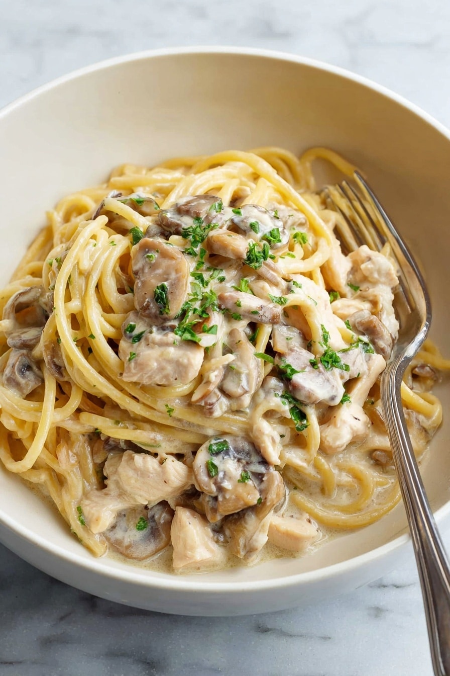 A white bowl holds creamy pasta with three main layers: at the bottom are light brown mushroom pieces coated in sauce, on top are thick, pale yellow spaghetti noodles mixed with tender white chicken pieces, all covered in a smooth, light cream sauce. Small green parsley bits are scattered on top, adding a touch of color. A silver fork rests inside the bowl on the right side. The bowl sits on a white marbled surface. Photo taken with an iphone --ar 2:3 --v 7 - Creamy Chicken Tetrazzini Bake, easy chicken casserole, cheesy pasta bake, weeknight dinner ideas, comforting chicken bake