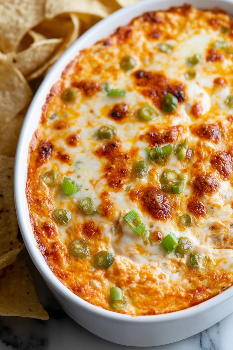 The image shows a close-up of a white oval dish filled with a baked cheesy dip. The top layer is golden brown melted cheese with small browned spots and melted orange cheese mixed in. Scattered on the surface are pieces of green onion adding a fresh green color contrast. The dish is placed on a white marbled surface, and in the background, there are some light and dark tortilla chips slightly out of focus. The melted cheese has a bubbly, slightly crispy texture on top photo taken with an iphone --ar 2:3 --v 7 - Buffalo Chicken Dip, spicy buffalo chicken appetizer, creamy hot wing dip, easy party dip, crowd-pleasing chicken dip