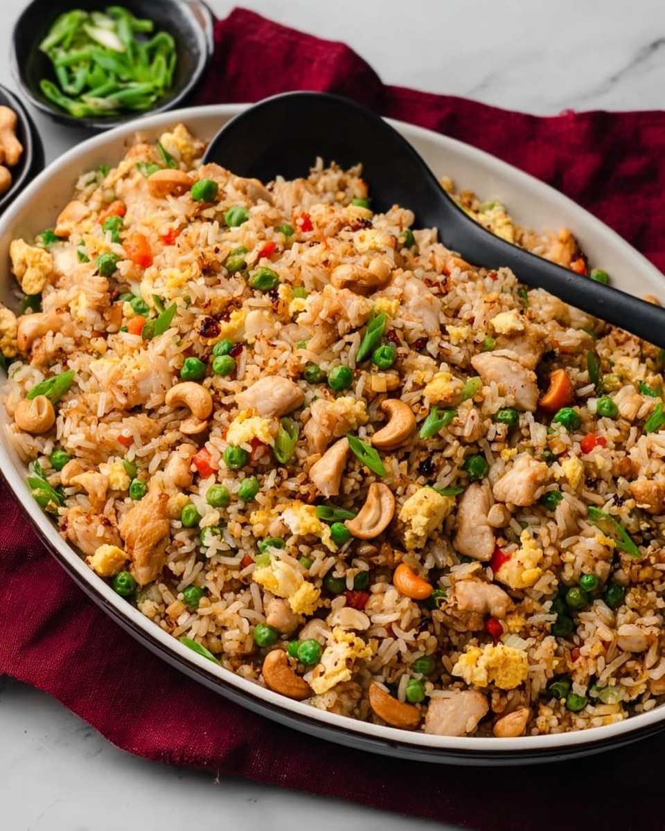 A large deep white oval dish is filled with a colorful fried rice dish with multiple layers of ingredients mixed together evenly. The base layer is light brown cooked rice, interspersed with small pieces of light golden scrambled eggs and light tan chunks of cooked chicken. Bright green sliced scallions and green peas are spread throughout, adding fresh color. There are also small orange carrot cubes and whole light brown cashew nuts, giving texture and visual contrast. A black spoon rests on the dish, ready to serve. The dish sits on a white marbled surface with a dark red cloth partially visible under the dish. photo taken with an iphone --ar 4:5 --v 7