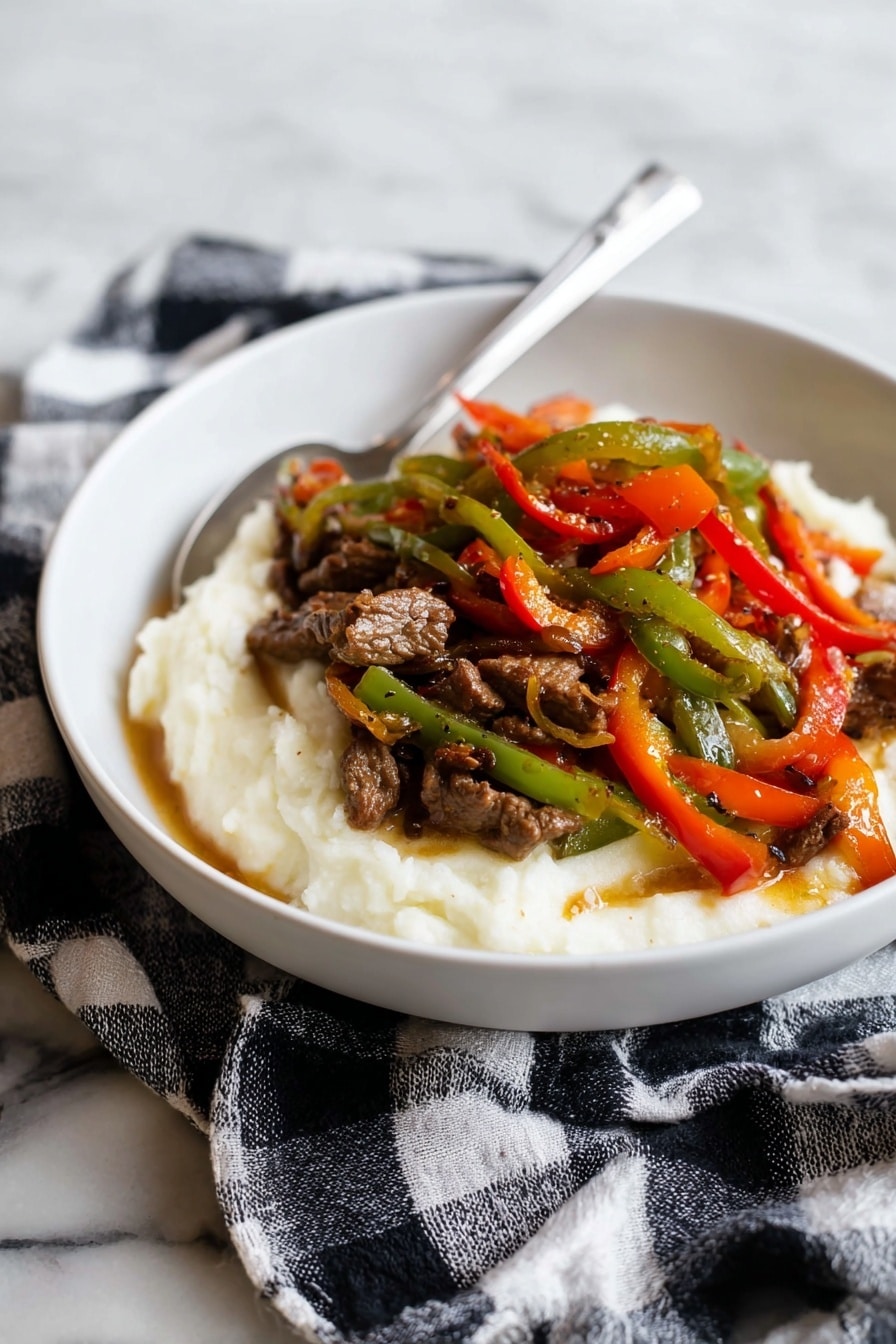 A white bowl holds a dish with two main layers: the bottom layer is a smooth, creamy white mashed texture, slightly spread around the edges. On top, there is a colorful mix of thin strips of cooked green, red, and orange bell peppers, mixed with small pieces of brown cooked meat, all glistening with a light sauce. A silver spoon rests inside the bowl, angled towards the back. The bowl sits on a black and white checkered cloth, placed on a white marbled surface. photo taken with an iphone --ar 2:3 --v 7 - Pepper Steak with Onion, pepper steak recipe, savory steak with peppers, hearty beef stir-fry, easy beef and onion dinner