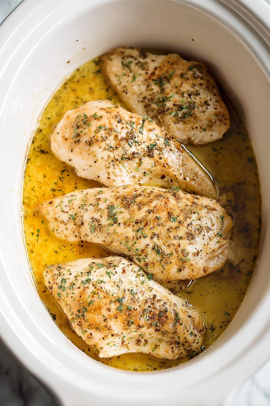 The image shows four pieces of cooked chicken placed side by side inside a white slow cooker. Each piece is light golden brown with some darker brown spots from cooking and sprinkled with green herbs and black pepper. The chicken sits in a shallow puddle of yellowish oil or broth that gives a slight shine on the surface. The inside of the slow cooker is white and smooth, contrasting with the warm tones of the chicken and liquid. The setting features a white marbled background. photo taken with an iphone --ar 2:3 --v 7 - Slow Cooker Juicy Seasoned Chicken Breast, flavorful chicken breasts, easy slow cooker chicken recipe, tender slow cooker chicken, healthy chicken dinner