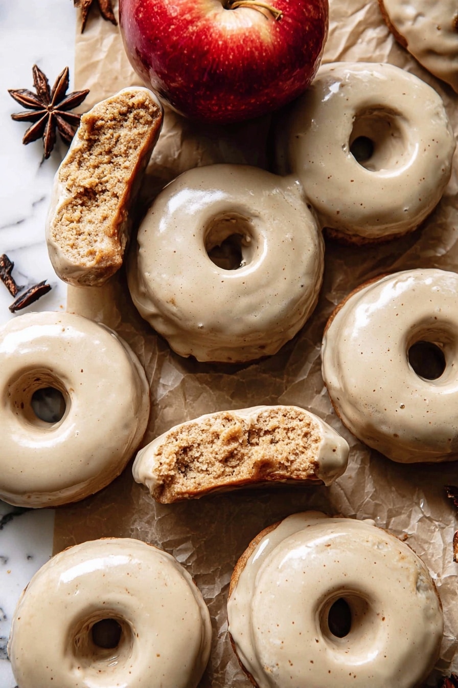 The image shows a group of doughnuts with a smooth, light beige glaze covering their top surfaces. Each doughnut has one round hole in the center and a slightly rough, crumbly texture inside where some are broken or bitten. The doughnuts are laid on a brown paper surface with pieces of white parchment paper scattered around. There is a shiny red apple near the center top of the image and a dark brown star anise near the upper left side. The background is a white marbled texture. photo taken with an iphone --ar 2:3 --v 7 - Baked Apple Cider Doughnuts with Cinnamon Maple Glaze, apple cider doughnuts, baked fall donuts, cinnamon maple glaze recipe, homemade apple donuts