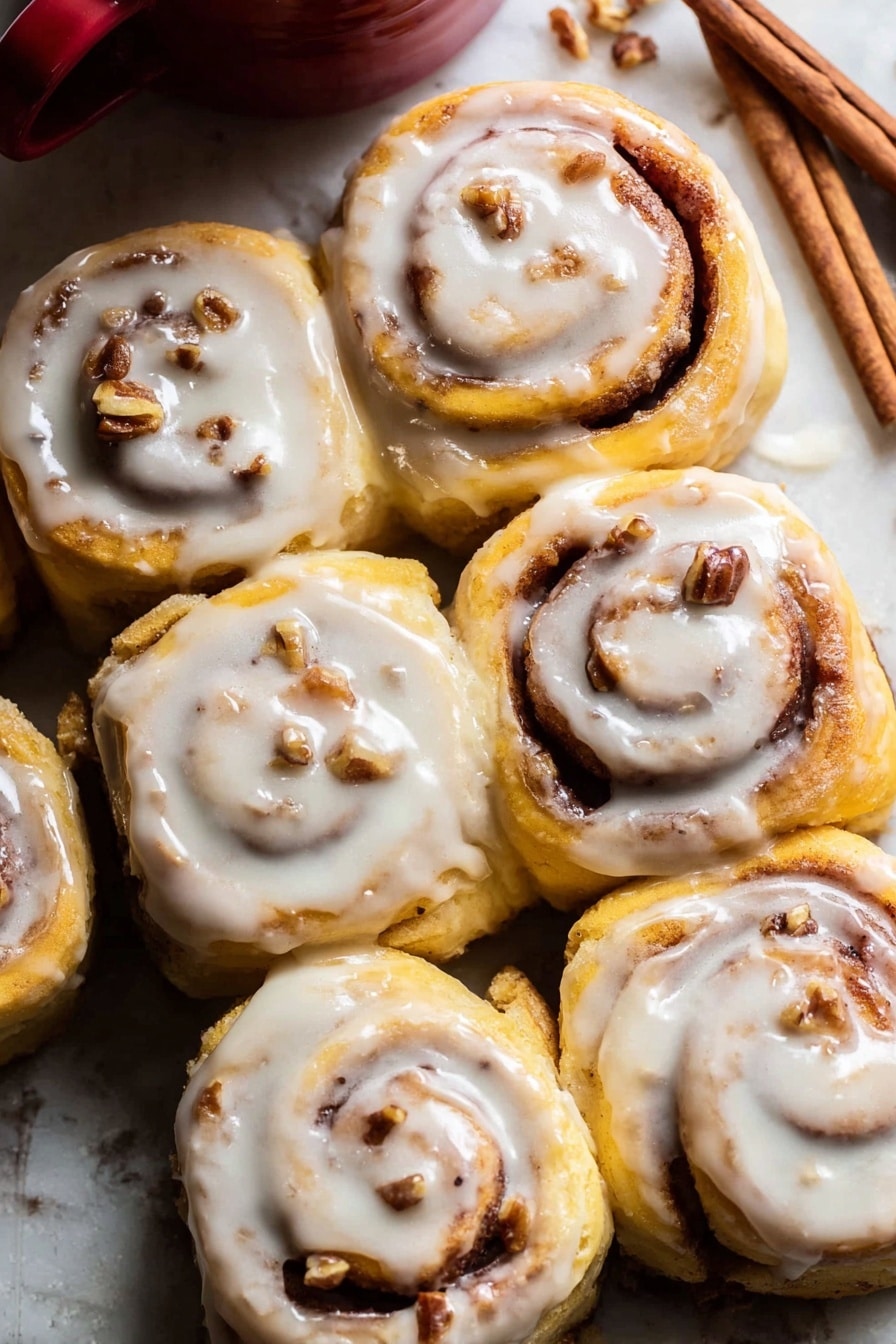 A close-up view of six cinnamon rolls arranged closely together on a white marbled surface. Each roll shows a soft golden-yellow dough spiraled with a dark cinnamon and sugar filling, with small pieces of nuts embedded within. A creamy white icing with a smooth texture generously covers the top of each cinnamon roll, slightly dripping down the sides. In the upper background, a few cinnamon sticks lay on the white marbled surface near the top right corner, next to a red ceramic cup partially visible. The warm tones of the rolls contrast nicely with the white icing and the white marbled surface, giving a fresh and inviting look. photo taken with an iphone --ar 2:3 --v 7 - Apple Cinnamon Rolls with Brown Butter Maple Icing, apple cinnamon rolls, brown butter maple frosting, apple cinnamon roll recipe, easy cinnamon roll with apple