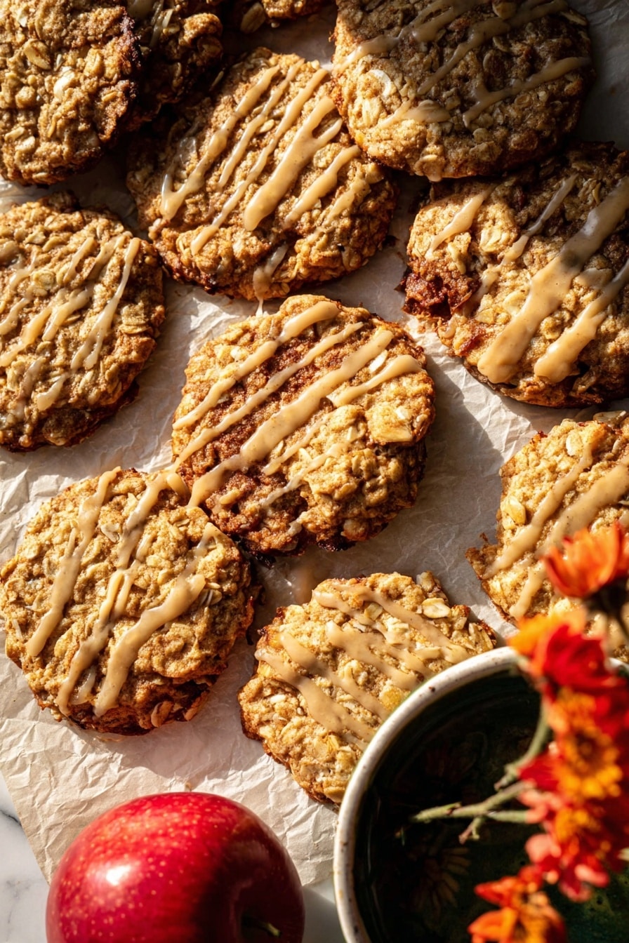 The image shows a close-up of several oatmeal cookies with a slightly rough texture from oats and nuts, arranged unevenly on parchment paper over a white marbled surface. Some cookies are stacked, while others lay flat, revealing their golden-brown baked edges with a few darker spots. A few cookies are drizzled with a light beige glaze in thin, irregular lines, adding shine and contrast to the surface. A bright red apple is partially visible in the bottom left, and the corner of a white bowl with a dark liquid and a yellow flower is seen in the top right, adding color variety. The warm lighting highlights the cookies' texture and the glaze’s glossiness. photo taken with an iphone --ar 2:3 --v 7 - Iced Apple Cider Caramel Oatmeal Cookies, fall-flavored oatmeal cookies, caramel apple cookies, cozy autumn cookie recipe, easy fall cookie ideas