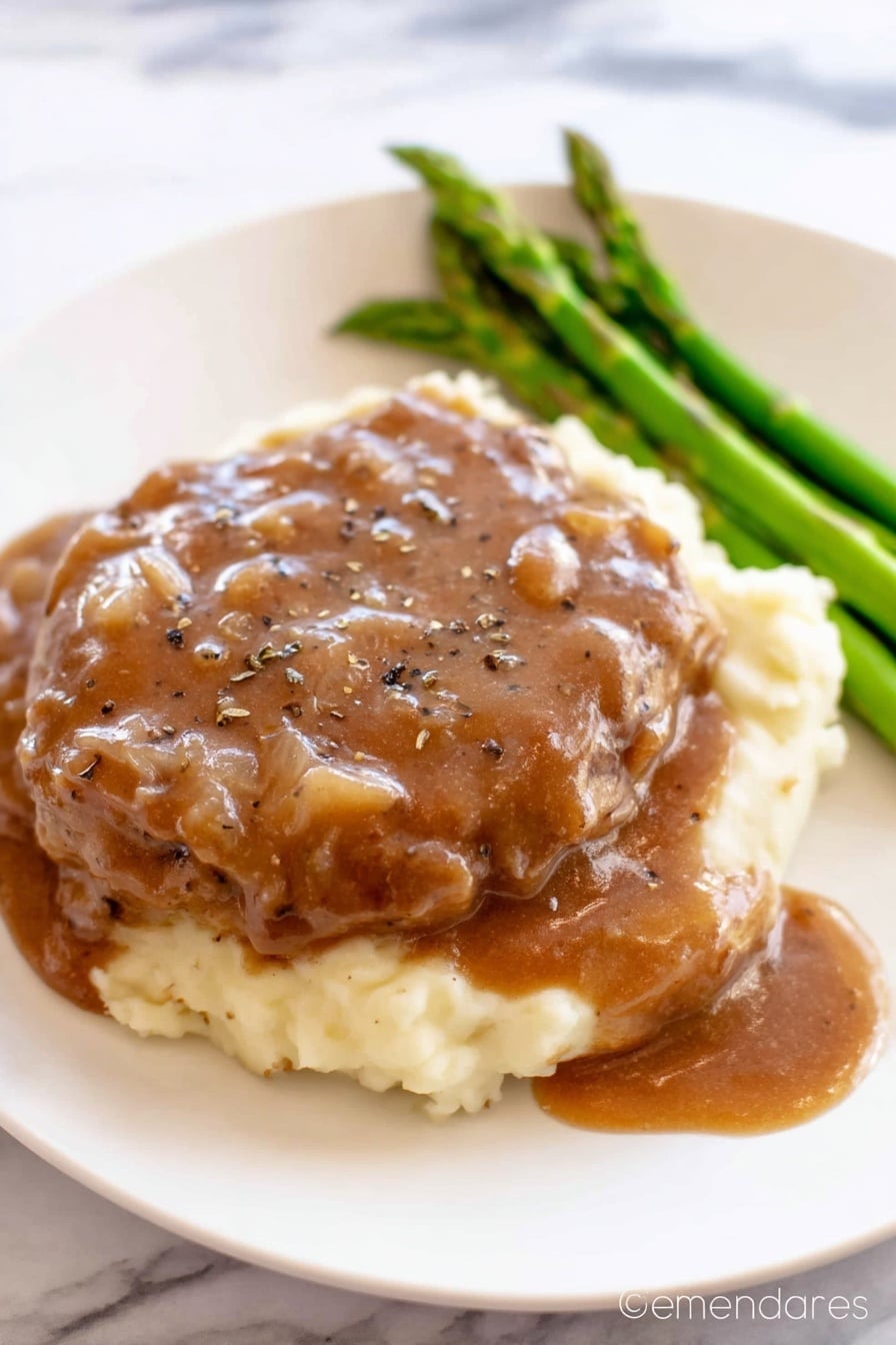 The image shows a white plate with three layers of food. The bottom layer is creamy mashed potatoes, which look soft and smooth in a pale cream color. On top of the mashed potatoes is a thick piece of cooked meat covered generously with a brown gravy sauce that has small onion bits and black pepper sprinkled on top. To the side of the meat, there is a small bunch of cooked green asparagus spears that add a fresh green color. The white plate sits on a white marbled surface. Photo taken with an iphone --ar 2:3 --v 7 - Crock Pot Cube Steak with Mushroom Gravy, slow cooker cube steak, easy mushroom gravy recipe, tender cube steak slow cooker, comfort food recipes