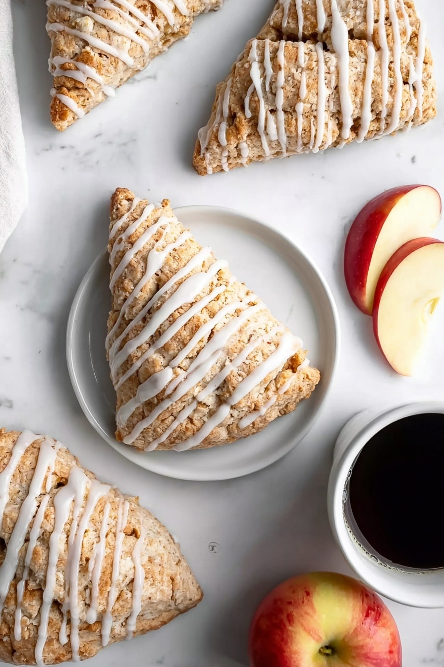 Several triangular scones are arranged on a white marbled surface, each covered in a light brown, crumbly texture with white icing drizzled in even stripes across the top. One scone sits on a simple white plate centered in the image, accompanied by two slices of red and yellow apple on the right side of the plate. Nearby, a whole apple with red and yellow skin is partly visible at the bottom right. In the upper right corner, a white cup filled with black coffee adds contrast. The scene is bright and clean, highlighting the soft texture of the scones and fresh fruit. photo taken with an iphone --ar 2:3 --v 7 - Apple Scones, easy apple scones, fall breakfast recipes, cinnamon apple baked goods, quick morning scones
