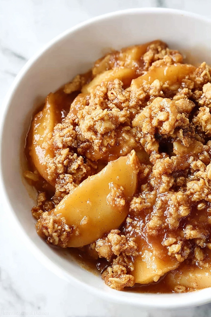 A white bowl holds a warm apple dessert with three distinct layers: the bottom layer has soft, golden-brown apple slices coated in syrupy juices; above this is a crumbly, textured topping made of oats and brown sugar that looks crunchy and slightly toasted; the top layer is a smooth, creamy white scoop of melted vanilla ice cream that softly spreads over the crumble. A silver spoon rests in the bowl, partially visible at the back, all placed on a white marbled surface. Photo taken with an iphone --ar 2:3 --v 7 - Healthy Apple Crisp with Oat-Pecan Topping, healthy apple crisp, gluten-free apple dessert, low-sugar apple crisp, nutritious fruit crisp