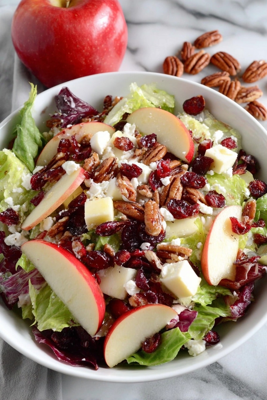 A white bowl filled with a fresh salad sits on a white marbled surface next to a red and yellow apple and a white towel with red stripes. The salad has a base layer of green leafy lettuce, topped with chunks of red and yellow apple pieces. Scattered over the apples are dried red cranberries and brown pecan nuts. The top layer is sprinkled with white crumbled cheese, adding a soft texture contrast to the crunchy ingredients. The photo taken with an iphone --ar 2:3 --v 7 - Honeycrisp Harvest Salad, autumn apple salad, maple bacon salad, apple cider vinaigrette salad, easy fall salad recipes