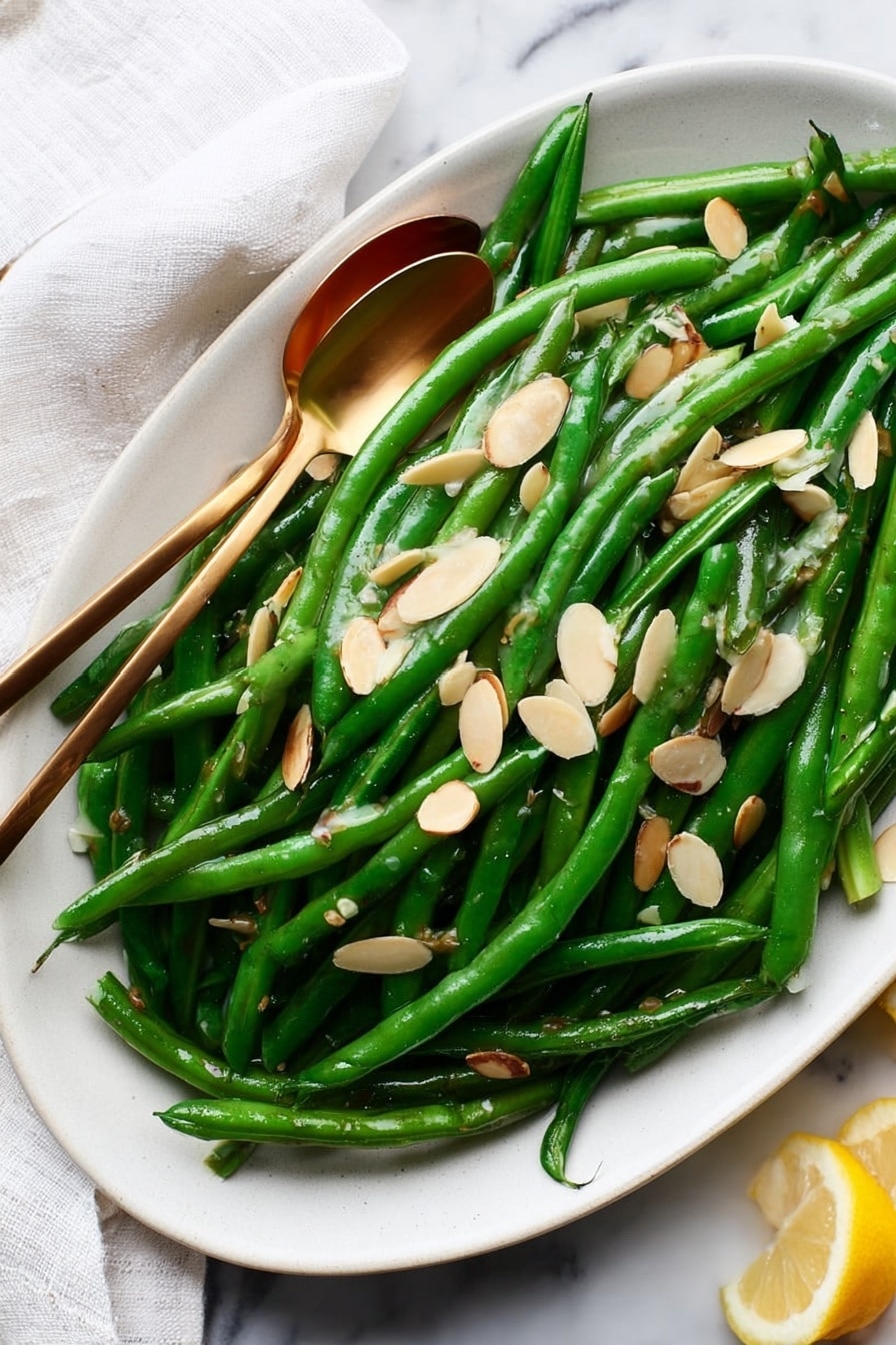 A white oval plate filled with bright green, shiny cooked green beans, arranged in a loose pile covering the plate. On top of the beans are thin, light tan almond slices scattered evenly, adding texture and contrast. Two small gold spoons rest on the beans, slightly overlapping, showing off the glossy surface of the veggies. The plate is placed on a white marbled surface, with a white cloth partially visible on the left side and a small piece of lemon on the right edge. The scene is well-lit, emphasizing the freshness and color of the dish. photo taken with an iphone --ar 2:3 --v 7 - Green Beans Almondine, easy green bean side dish, almond green beans, lemon green beans recipe, quick vegetable side