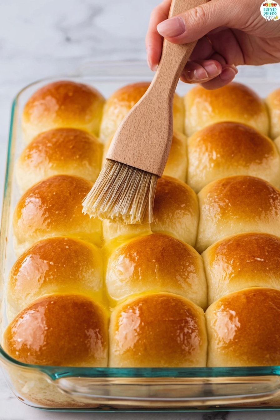 A glass baking dish holds a layer of 12 evenly arranged soft dinner rolls, each roll golden brown on top and shiny from being brushed with melted butter. The rolls sit closely together in a grid pattern with a soft, slightly fluffy texture. A woman's hand holds a light wooden brush with brown bristles, applying butter to the top of one roll. The background is a white marbled surface. photo taken with an iphone --ar 2:3 --v 7 - Homemade Fluffy Dinner Rolls, soft dinner roll recipe, buttery dinner rolls, easy bread rolls, homemade bread rolls