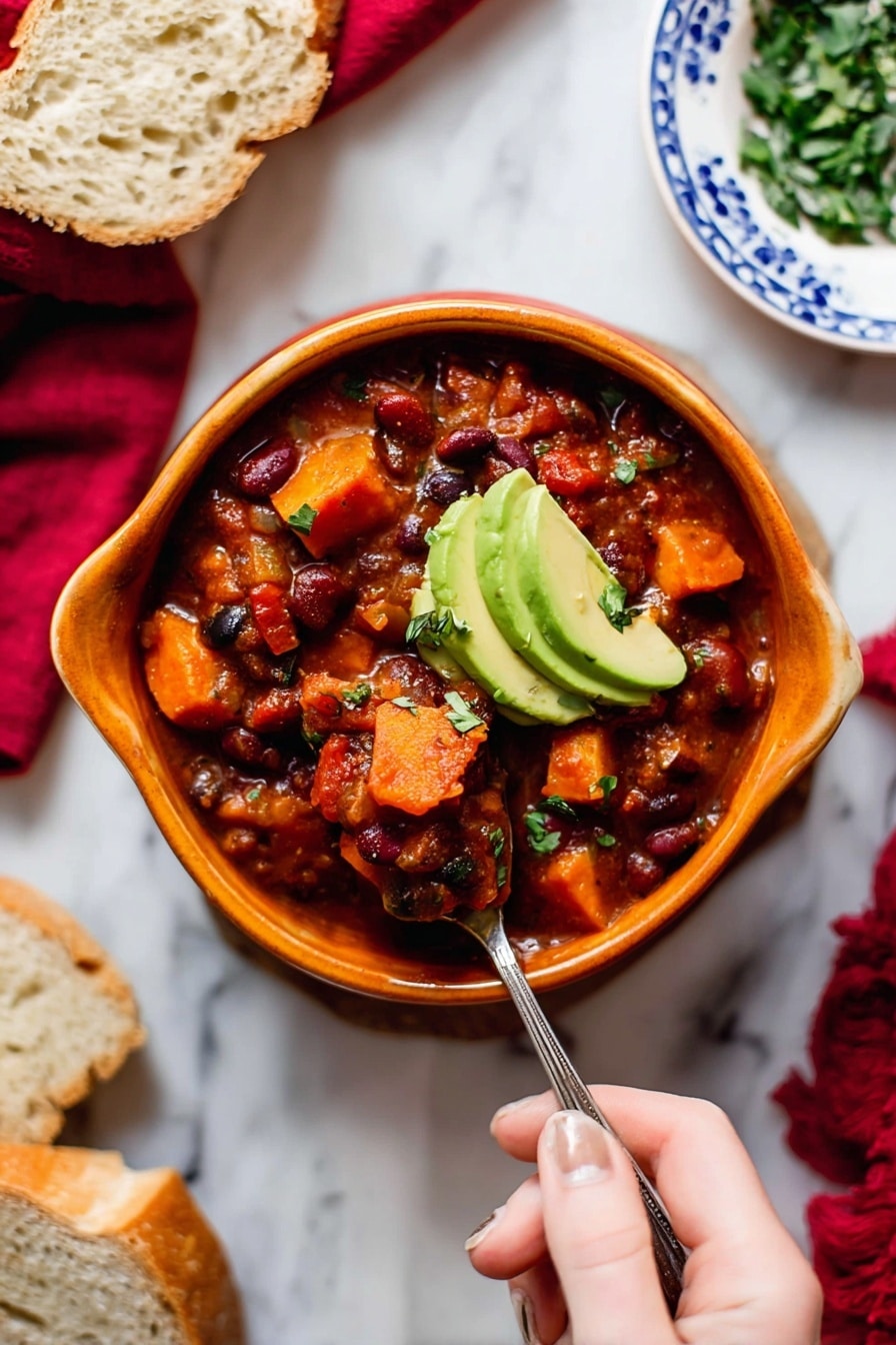 A bowl of thick chili with visible layers of chunky tomato sauce, red and black beans, and soft, cubed orange sweet potatoes, topped with two slices of green avocado. The chili is served in a curved, orange bowl on a white marbled surface, with a woman's hand holding a spoon scooping into the chili. Surrounding the bowl are slices of bread on a white plate with blue patterns, a small bowl of chopped green herbs, and a red cloth in the background. Photo taken with an iphone --ar 2:3 --v 7 - Vegetarian Pumpkin Chili, hearty pumpkin chili, healthy vegetarian chili, easy pumpkin chili recipe, cozy fall chili