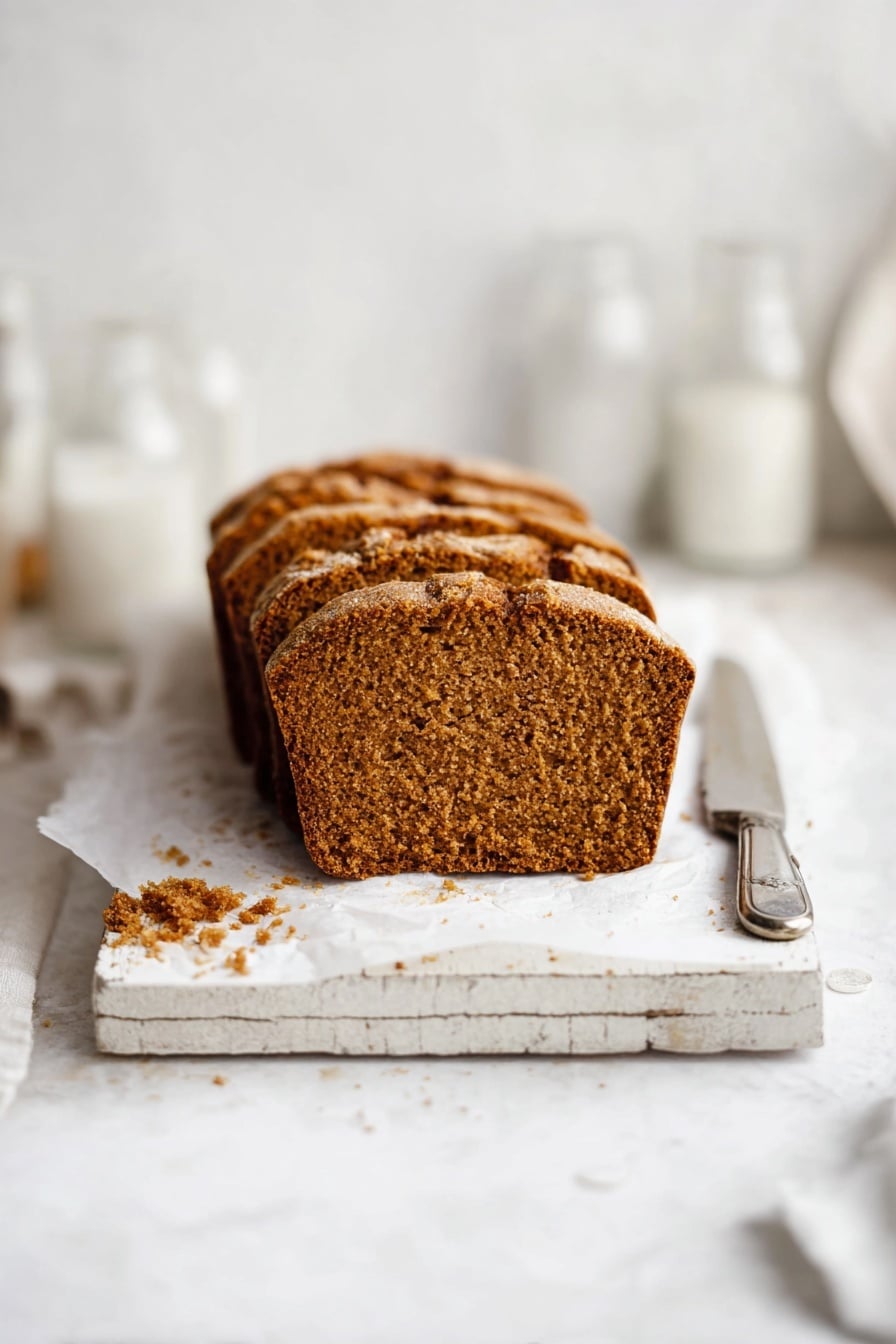 The image shows a loaf of brown bread sliced into six pieces, placed on white parchment paper on a rustic white wooden board. The bread has a golden-brown crust with a slightly rough texture and a dense, soft interior with small air holes. There are some crumbs scattered around the bread on the white marbled surface. To the upper right of the bread, there is a silver knife resting on top of a folded piece of white paper with text, and in the upper left, a clear empty glass and a white container are partially visible. Photo taken with an iphone --ar 2:3 --v 7 - Easy Moist Pumpkin Bread, fall pumpkin bread, moist pumpkin bread recipe, pumpkin bread with spices, soft pumpkin quick bread