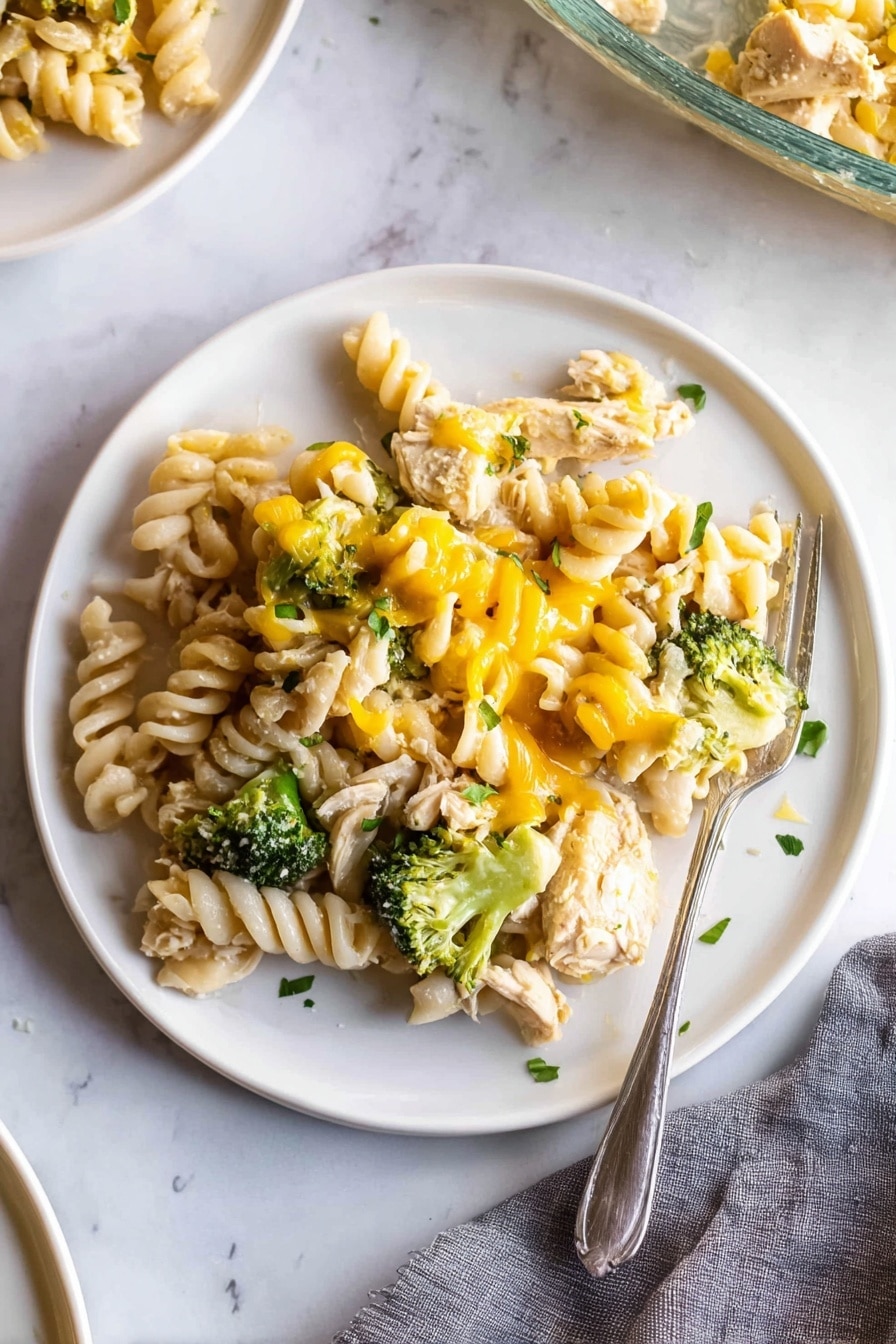 A white plate on a white marbled surface holds a serving of rotini pasta mixed with chunks of cooked chicken, bright green broccoli florets, and melted yellow cheese drizzled unevenly over the top. The pasta spirals are pale cream and slightly glossy, nestled closely with tender pieces of chicken and small green broccoli heads, all lightly sprinkled with small bits of fresh green herbs. A silver fork rests partially on the right edge of the plate, with its tines touching some pasta. There is a gray cloth napkin in the lower right corner and a partial view of another white plate and a glass baking dish in the background. Photo taken with an iphone --ar 2:3 --v 7 - Easy Chicken Noodle Casserole, chicken noodle casserole, cheesy chicken casserole, creamy chicken noodle bake, simple chicken casserole