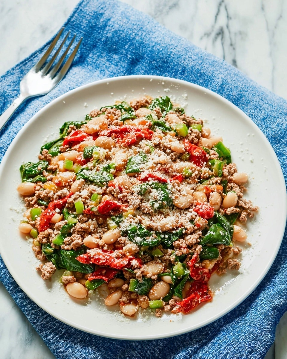 The dish is a colorful mix served on a white plate placed on a white marbled surface, with a blue napkin and a fork beside it. The food looks like a layered salad or stir fry with four main layers: small grains of light brown cooked ground meat form the first base layer, mixed evenly with plump white beans; bright green spinach leaves and diced green bell peppers are scattered through the mix, adding fresh texture; chunks of red tomatoes or roasted red peppers create spots of bright color on top; the dish is sprinkled lightly with a powdery white cheese or seasoning. The whole dish has a warm, hearty look and a fresh, healthy vibe. photo taken with an iphone --ar 4:5 --v 7