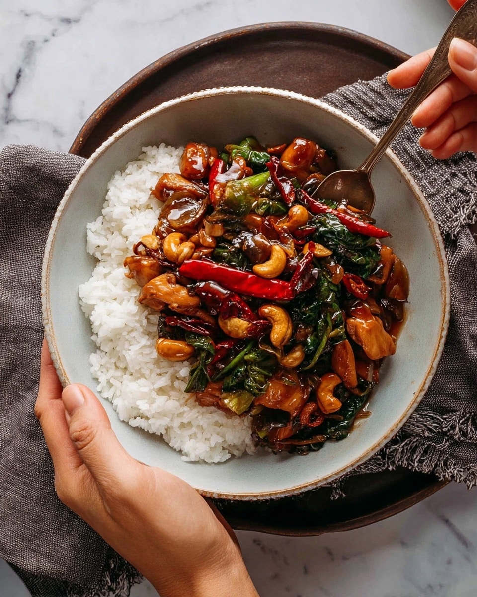 A bowl filled with three layers is shown. The bottom layer is white steamed rice, fluffy and soft. On top of the rice, there is a vibrant brown stir-fry with pieces of cooked chicken, shiny dark green leafy vegetables, whole golden brown cashews, slices of red chili peppers, and light brown onion slices, covered in a glossy sauce. A woman's hand holds the white bowl from the left side, while another woman's hand holds a spoon stirring the food on the right side. The bowl sits on a dark brown plate with a rough gray cloth beneath it, all set against a white marbled surface. Photo taken with an iphone --ar 4:5 --v 7