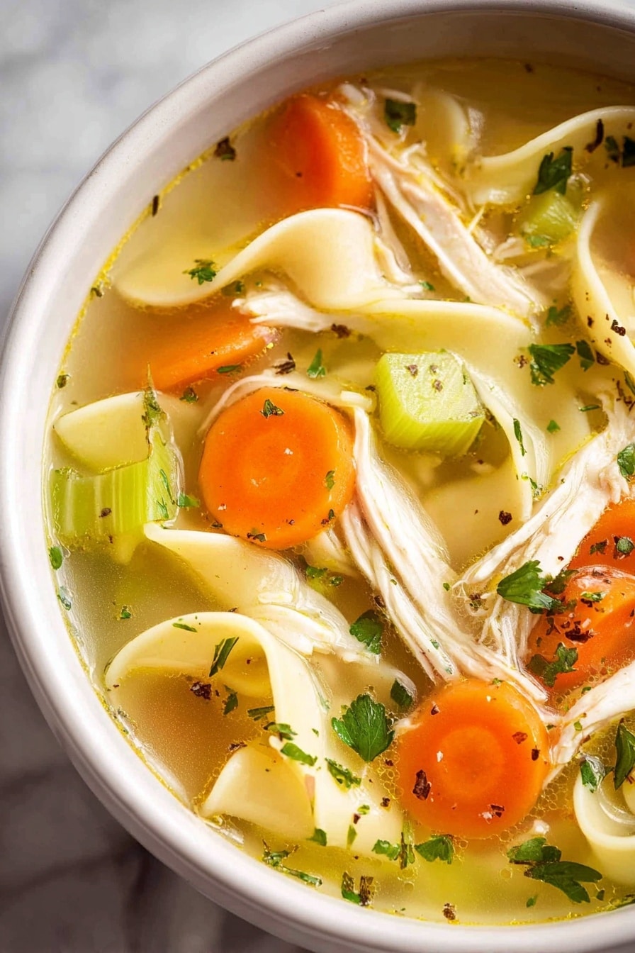 A white bowl filled with clear golden broth containing long, flat yellow noodles, small orange carrot cubes, and green celery pieces. Shredded light beige chicken is spread evenly throughout, with flecks of green herbs and black pepper for extra color. A single dark brown bay leaf is visible near the bottom, and a silver spoon is partially submerged in the soup. The bowl rests on a white marbled surface with a small white cup of chopped green herbs and another white cup with a white creamy substance in the background, photo taken with an iphone --ar 2:3 --v 7 - Homemade Chicken Noodle Soup from Scratch, homemade chicken noodle soup, scratch-made chicken soup, homemade chicken soup recipe, comforting chicken noodle soup