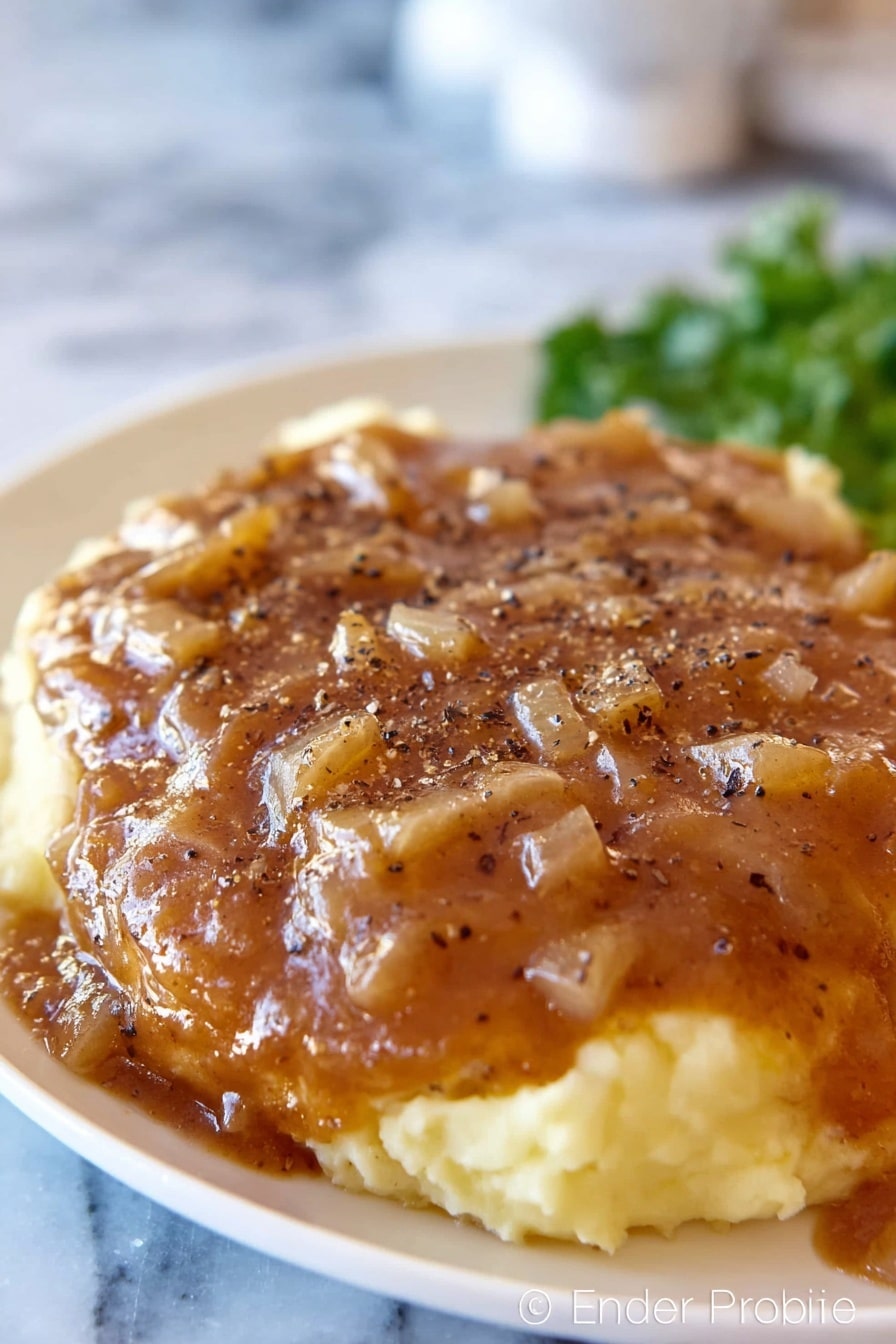 A white plate with mashed potatoes as the base layer, creamy and light yellow in color with a smooth texture, topped with a thick brown gravy that has small diced onion pieces spread evenly across the surface. The gravy covers a large piece of meat underneath, sprinkled with black pepper flakes. The background shows an out-of-focus white marbled surface and some green garnish in the back. Photo taken with an iphone --ar 2:3 --v 7 - Crock Pot Cube Steak with Mushroom Gravy, slow cooker cube steak, easy mushroom gravy recipe, tender cube steak slow cooker, comfort food recipes