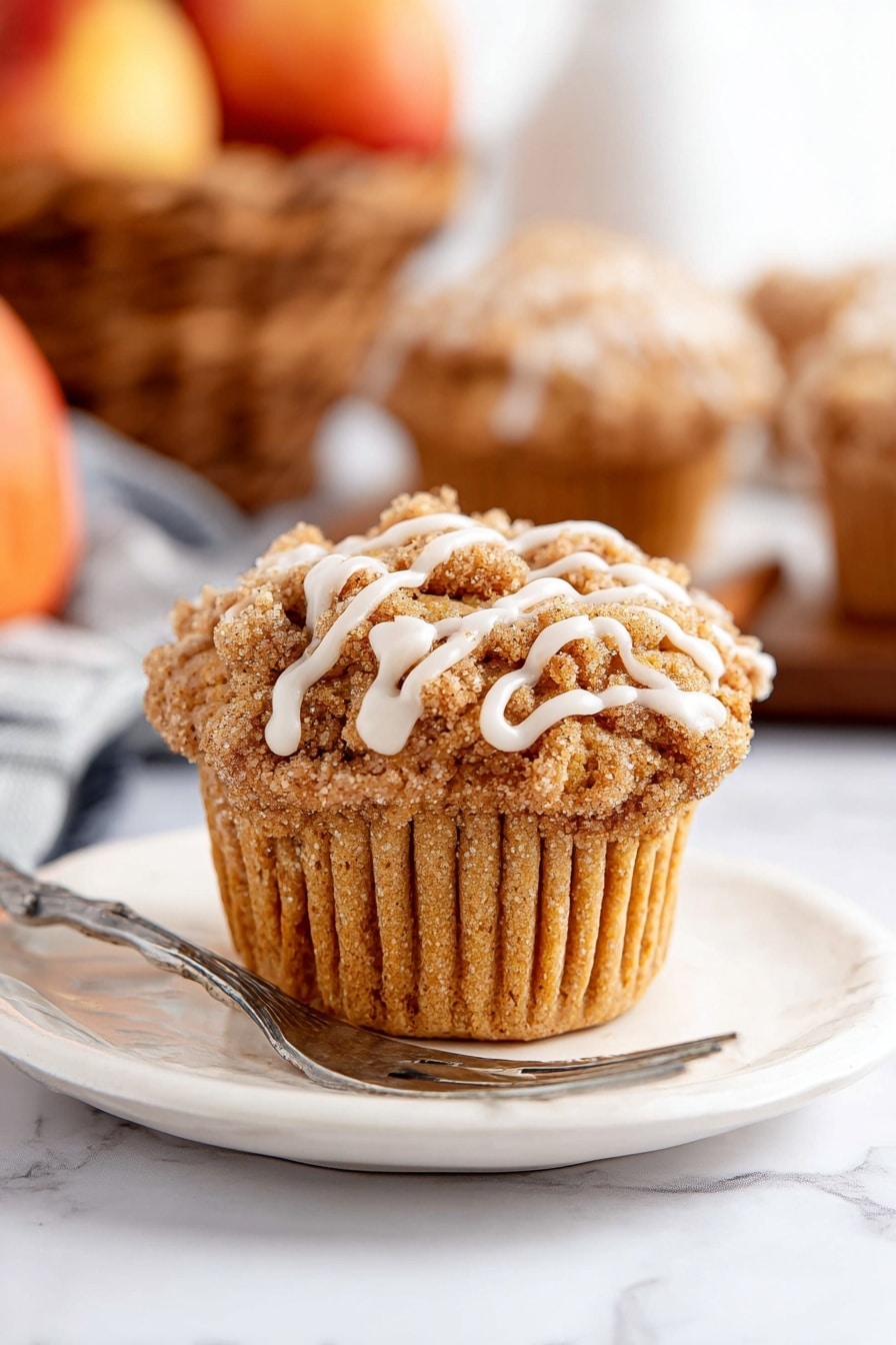 A single muffin sits centered on a white plate with a silver fork resting next to it. The muffin has a light brown, soft textured base with a crumbly streusel topping in a darker brown shade. White icing drizzle runs in thin lines over the streusel, adding contrast. The plate and muffin are placed on a white marbled surface. In the background, blurred muffins and a basket of apples add warm tones but stay out of focus. Photo taken with an iphone --ar 2:3 --v 7 - Cinnamon Apple Muffins with Crumble Topping and Glaze, apple cinnamon muffins, homemade apple muffins, fall breakfast muffins, easy apple muffin recipe