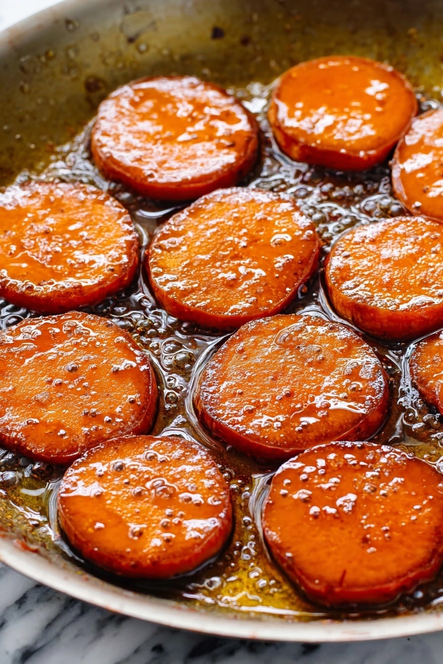 The image shows a white oval dish filled with thick, round slices of cooked sweet potatoes. The sweet potato slices are covered in a shiny, dark caramel-like sauce that makes them look glossy and soft. The slices are arranged closely together, slightly overlapping, and their orange color is rich and warm, enhanced by the dark syrup. The sauce pools around the edges of the slices, filling the bottom of the dish, and the texture looks smooth and sticky. The setting is on a white marbled surface. photo taken with an iphone --ar 2:3 --v 7 - Sweet Potatoes with Bourbon Glaze, bourbon glazed sweet potatoes, holiday side dish recipes, caramelized sweet potatoes, easy festive side dishes