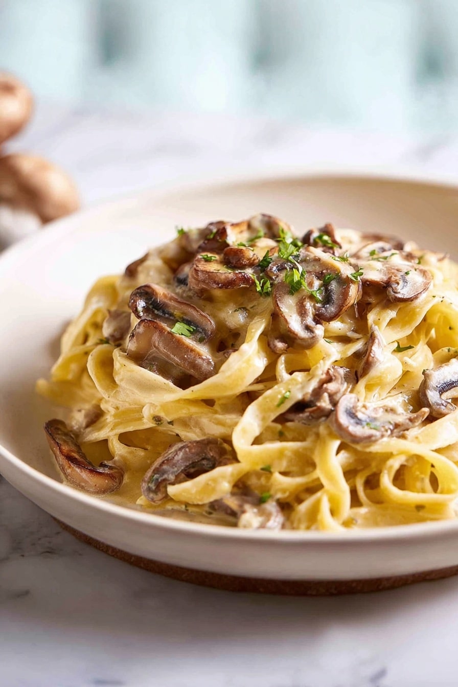 The image shows a mound of creamy fettuccine pasta on a plain white plate. The pasta is light yellow and looks soft and smooth, with thin sauce coating each strand. On top and mixed through are brown sliced mushrooms with a slight gloss, adding texture and earthy colors. Small green herb bits are sprinkled over the pasta, giving little pops of color. The plate sits on a white marbled surface, with a blurred background that is softly lit showing a light blue and white shade. Photo taken with an iphone --ar 2:3 --v 7 - Creamy Mushroom Pasta, easy mushroom pasta, quick mushroom pasta dinner, homemade mushroom pasta, comfort food mushroom pasta