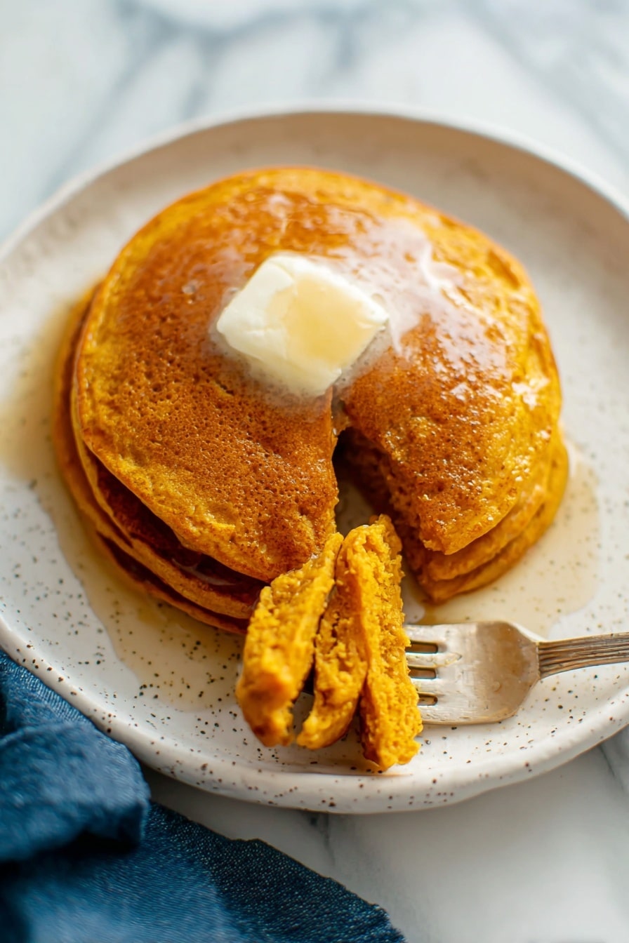 There is a white plate with three golden-brown pancakes stacked slightly unevenly with a clear textured top pancake showing tiny holes. A gold fork rests on the top edge of the plate. Next to the plate on the white marbled surface, there is a pile of more pancakes on another white plate that shows their soft, porous texture. A small white jug with dark syrup is near the plates, and a blue cloth is placed on the surface beside the plate. The photo taken with an iphone --ar 2:3 --v 7 - Whole Wheat Pumpkin Pancakes, Pumpkin Pancake Recipes, Healthy Pumpkin Breakfast, Fall Flavored Pancakes, Wholesome Breakfast Ideas
