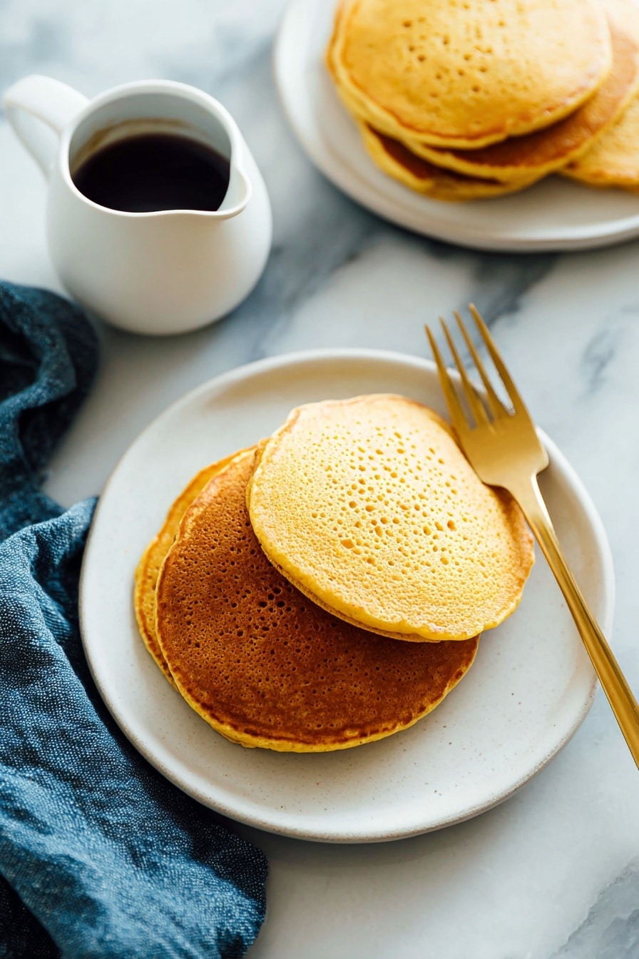 There is a stack of three golden-brown pancakes on a white plate. The pancakes have a slightly porous texture and are arranged with the largest at the bottom and the smallest on top. A light amber syrup is poured over the top pancake, dripping down the sides and pooling a little on the plate. A silver fork rests under the stack, on the left side, and the plate sits on a white marbled surface with a blue cloth nearby. The lighting is soft and natural, highlighting the warm tones of the pancakes and syrup. photo taken with an iphone --ar 2:3 --v 7 - Whole Wheat Pumpkin Pancakes, Pumpkin Pancake Recipes, Healthy Pumpkin Breakfast, Fall Flavored Pancakes, Wholesome Breakfast Ideas