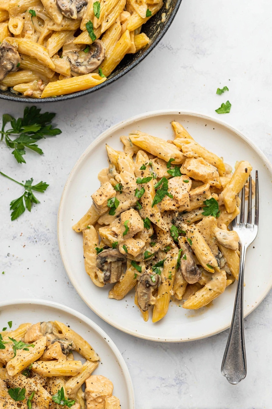 A white plate holds creamy pasta made of penne noodles mixed with pieces of cooked chicken and sliced mushrooms. The pasta and chicken are coated with a light beige creamy sauce, giving a smooth texture. The mushrooms are brown with soft white inside visible, placed evenly throughout. Green parsley leaves are sprinkled on top, adding a fresh color contrast, and there are small bits of red pepper flakes and black pepper scattered over the dish. A silver fork is partly stuck into the pasta on the right side, resting on the plate. The whole scene is set on a white marbled surface, creating a clean and bright background. photo taken with an iphone --ar 2:3 --v 7 - Creamy Chicken Marsala Pasta, Chicken Marsala pasta, creamy chicken pasta, mushroom marsala sauce, easy chicken pasta recipes