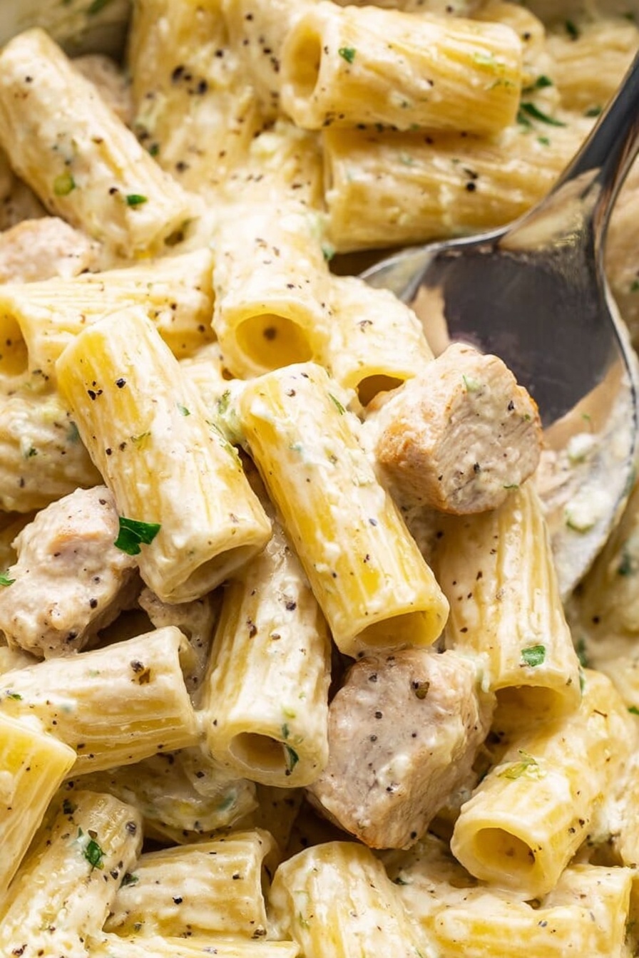 This close-up image shows tubes of rigatoni pasta covered in a creamy white sauce, mixed with small pieces of cooked chicken that have a light brown sear. The pasta is speckled with green herbs and some black pepper bits, and has a smooth, slightly glossy texture. The sauce looks thick and coats every piece evenly. A shiny metal spoon is partially visible picking up some pasta and chicken on the right side. The background is a white marbled surface. Photo taken with an iphone --ar 2:3 --v 7 - Creamy Lemon Chicken Pasta, Lemon Chicken Pasta, Easy Pasta Dinner, Quick Chicken Pasta, Zesty Creamy Pasta