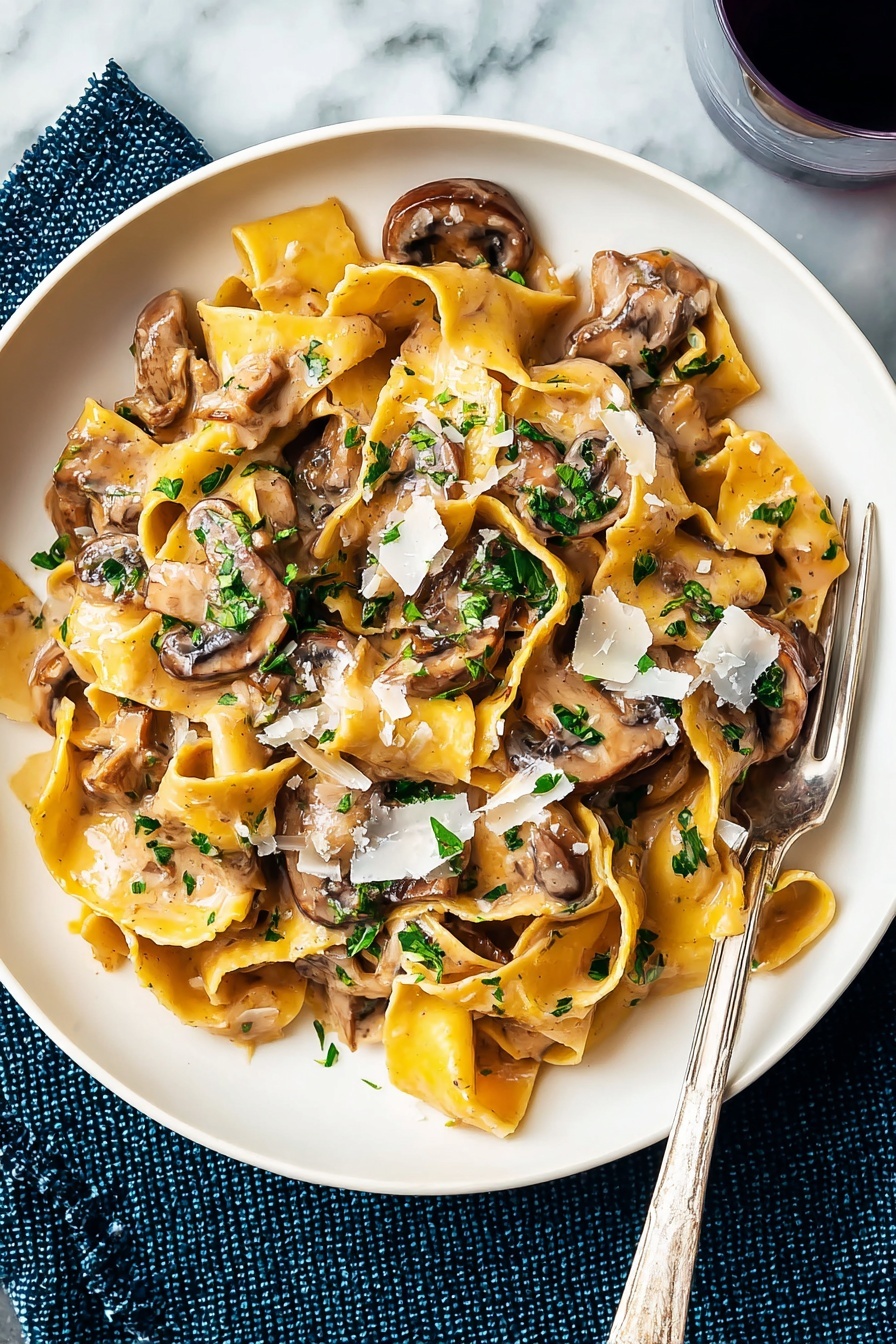 A white plate holds a serving of wide, yellow pasta ribbons cooked in a light brown creamy mushroom sauce. On top, there are several brown, slightly browned mushroom slices mixed throughout the pasta, adding a smooth texture. Bright green chopped herbs are sprinkled over the dish, giving a fresh contrast. Light shavings of white cheese are scattered on top, melting slightly into the sauce. A silver fork rests on the edge of the plate, and the plate is placed on a white marbled surface with a dark blue textured cloth beneath one side. Photo taken with an iphone --ar 2:3 --v 7 - Vegetarian Mushroom Stroganoff, mushroom stroganoff recipe, vegan mushroom stroganoff, creamy mushroom pasta, meat-free comfort food
