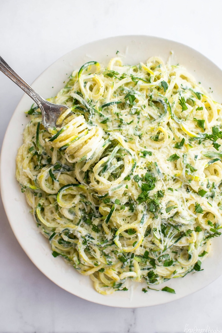 A white plate filled with a creamy dish made of spiral-shaped green and light yellow vegetable noodles, mixed with a white creamy sauce that coats the noodles evenly. There are small green herb leaves sprinkled throughout the dish, adding a fresh look. A silver fork is twirling some of the noodles in the top left area of the plate. The plate rests on a white marbled surface. photo taken with an iphone --ar 2:3 --v 7 - Zucchini Alfredo, Low-Carb Dinner, Healthy Pasta Alternatives, Gluten-Free Zucchini Noodles, Creamy Vegetable Alfredo