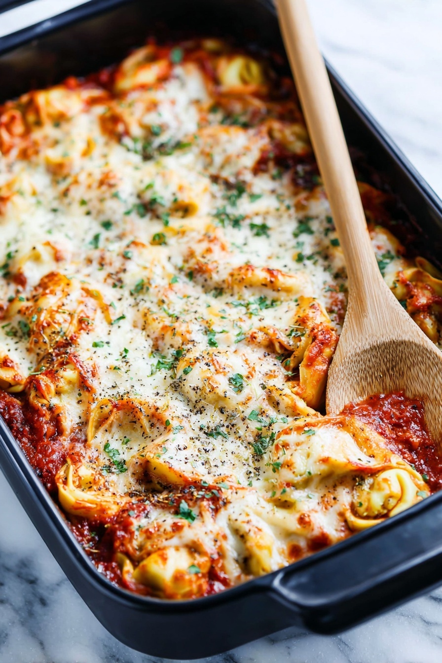 A black baking dish holds a layered tortellini pasta bake with a rich tomato sauce. The top layer is melted white cheese, bubbly and slightly browned in spots, mixed with twisted yellow tortellini filled pasta visible underneath. The sauce is deep red with bits of herbs and small chunks of meat mixed in. Scattered green herb leaves add color contrast across the top. The dish rests on a white marbled surface with a folded striped cloth nearby and a wooden-handled black spoon on the right edge of the frame, creating a cozy kitchen feel. photo taken with an iphone --ar 2:3 --v 7 - Cheesy Baked Tortellini Casserole, cheesy tortellini casserole, baked tortellini recipes, comforting pasta dishes, easy baked pasta