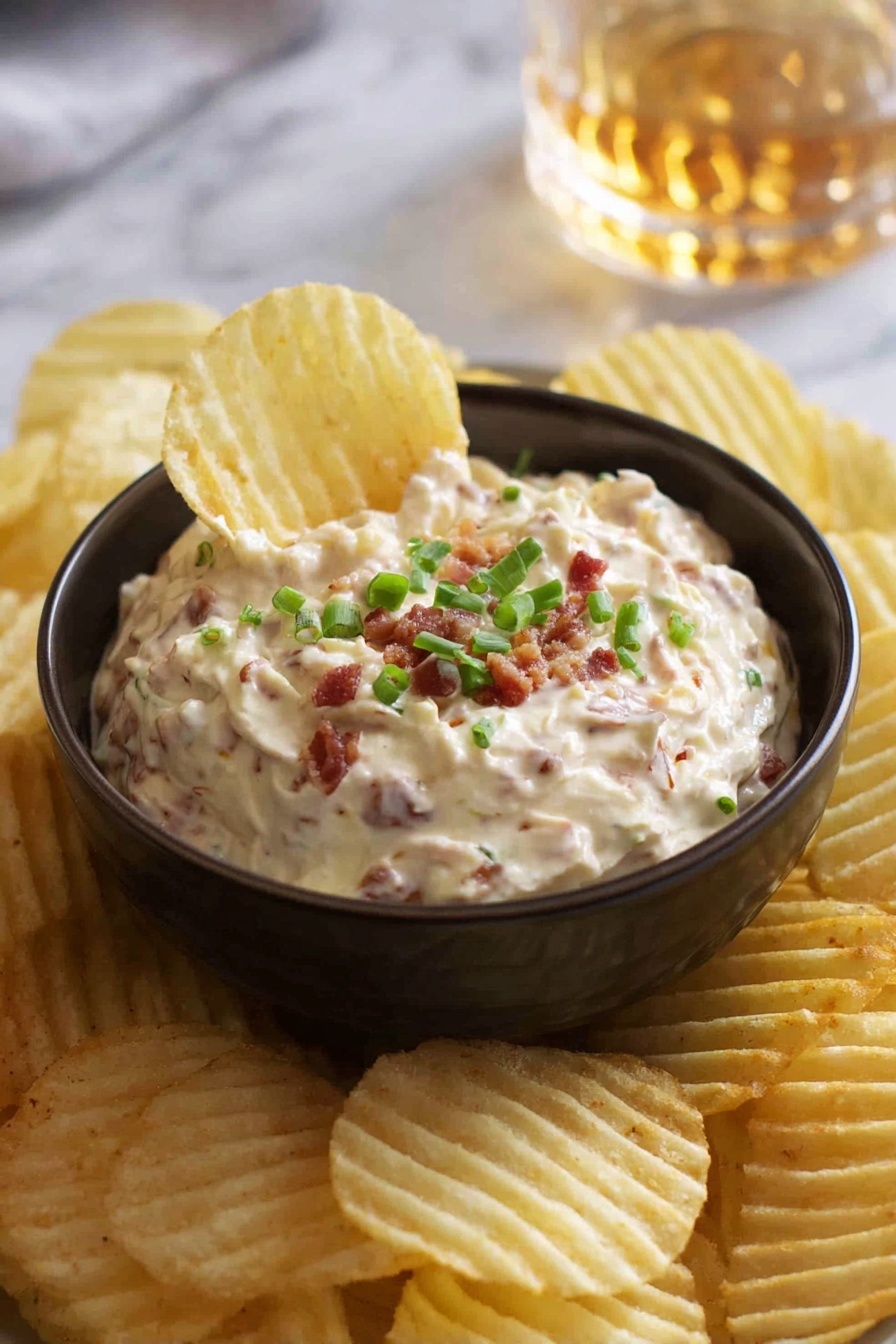 A small dark bowl filled with creamy dip that has small pieces of bacon mixed in, topped with chopped green onions. The bowl sits on a white marbled surface and is surrounded by many ridged potato chips, one chip is dipped into the bowl showing the texture of the dip. In the background, there is a blurred glass with a golden liquid. Photo taken with an iphone --ar 2:3 --v 7 - Homemade French Onion Dip, easy French onion dip, creamy onion dip recipe, homemade party dip, savory onion dip for snacks