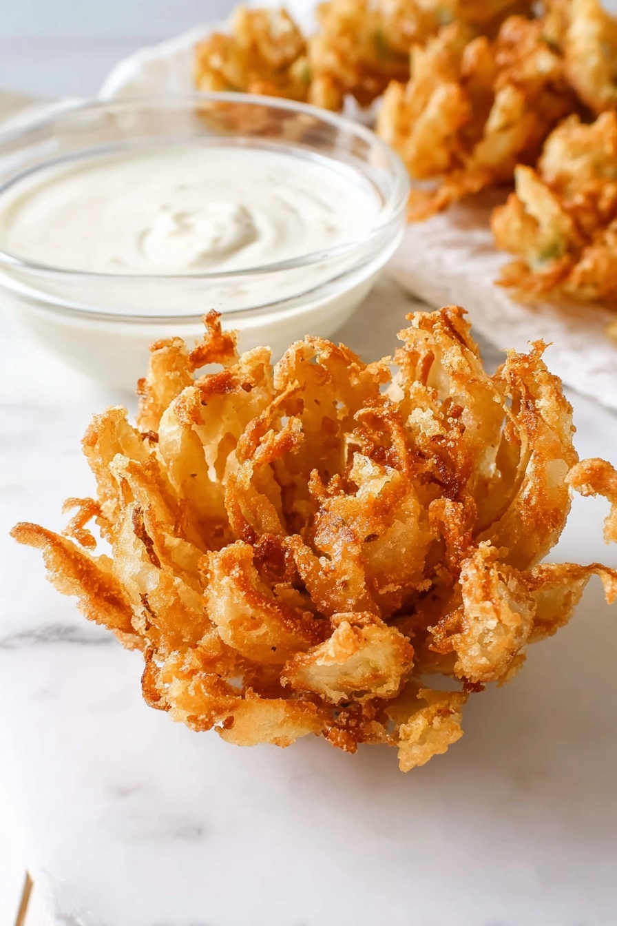A close-up image of a golden-brown, crispy fried onion blossom placed on a white marbled surface. The onion blossom is fully opened like a flower with many uneven, crunchy petals showing a textured, fried batter coating. Next to it is a small clear glass bowl filled with smooth, creamy white dipping sauce. In the background, some more fried onion blossoms are slightly blurred. The scene is bright and simple with clean white tones and a soft shadow under the food. photo taken with an iphone --ar 2:3 --v 7 - Homemade Blooming Onion, Blooming Onion with Dipping Sauce, Crispy Blooming Onion recipe, How to make blooming onion, Best blooming onion at home