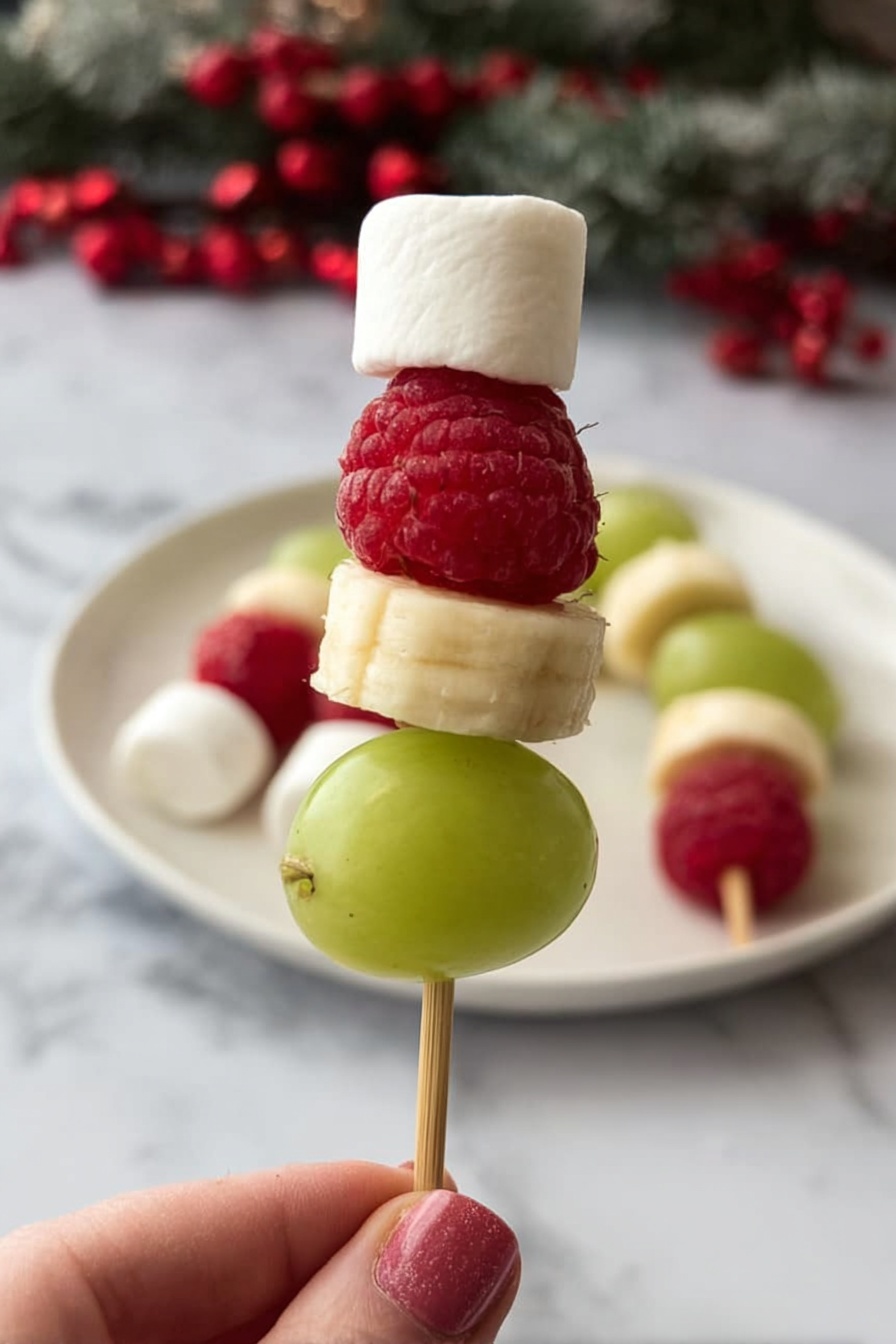 A small skewer held by a woman's hand shows four layers: at the bottom, a smooth, green grape; above it, a round, pale yellow banana slice; next a bright red raspberry with visible bumpy texture; and on top, a small white marshmallow shaped like a cube. In the background, more similar skewers rest on a white plate on a white marbled surface with some red berries. The overall look is fresh, colorful, and festive. photo taken with an iphone --ar 2:3 --v 7 - Grinch Fruit Kabobs, healthy holiday snacks, festive fruit skewers, kids friendly Christmas treats, colorful holiday party ideas