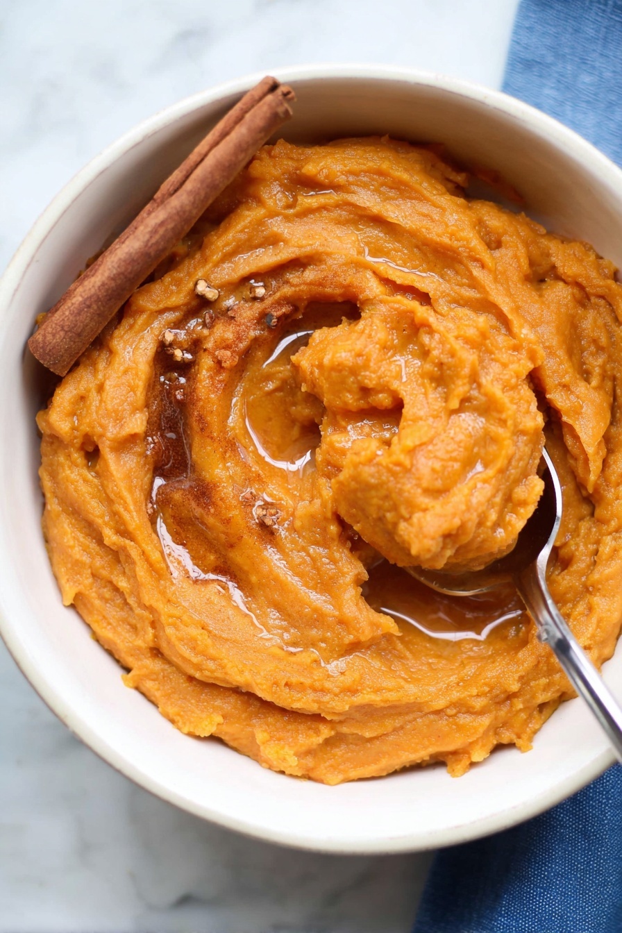 The image shows a white bowl filled with thick, smooth orange sweet potato mash. The mash has swirled texture with a shiny surface, topped with a light drizzle of syrup pooling slightly in the center. A cinnamon stick rests on the left side of the mash, and small bits of brown spices are sprinkled on top. A metal spoon is scooping up part of the mash on the right side, with the spoon handle extending out of the bowl. The bowl sits on a white marbled surface with a bit of a blue cloth visible in the bottom right corner. Photo taken with an iphone --ar 2:3 --v 7 - Brown Butter Mashed Sweet Potatoes, sweet potato side dish, cozy holiday side, roasted sweet potato mash, nutty flavored sweet potatoes