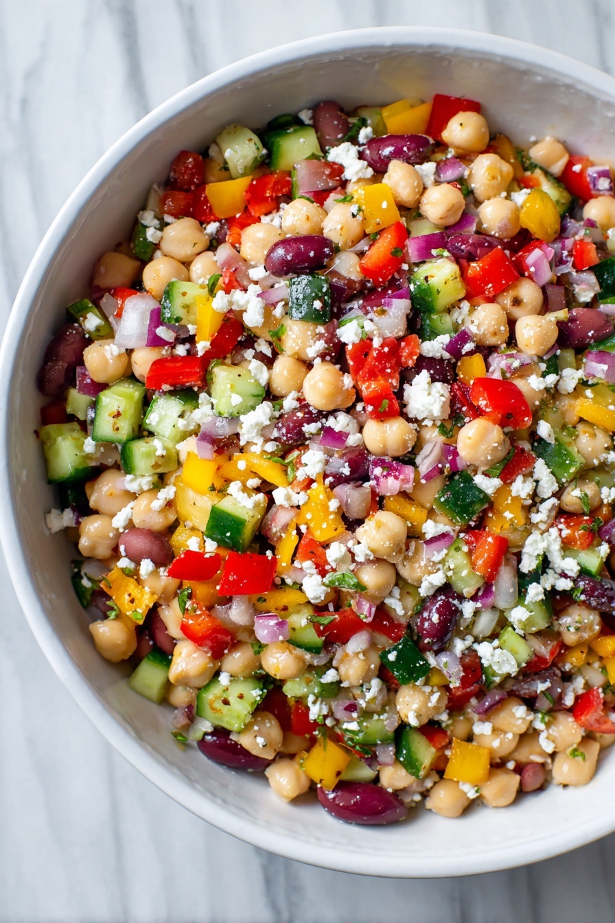 A white bowl filled with a colorful bean salad showing multiple layers of small, chopped vegetables and legumes. The base layer is made of light beige chickpeas and white beans mixed with a scattering of deep purple olive slices. On top, there is a thick mix of small diced bright red bell peppers, yellow bell peppers, green cucumbers, and red onions, creating a vibrant patchwork of colors. White crumbled cheese is sprinkled evenly over the salad, adding a soft, crumbly texture. The surface of the salad looks fresh, moist, and lightly coated in dressing. The bowl rests on a white marbled texture. Photo taken with an iphone --ar 2:3 --v 7 - Creamy Chickpea and Bean Salad, healthy bean salad, protein-packed vegetarian salad, quick lunch salad, refreshing veggie salad