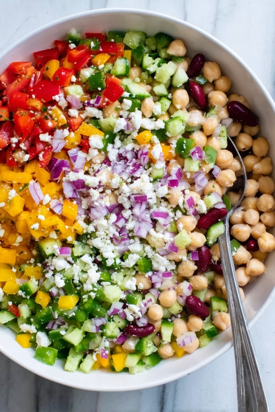 The image shows a white bowl filled with a colorful salad made of many small chopped vegetables and beans. There are five visible layers: at the bottom is a mix of small round chickpeas and white beans with a creamy texture, above that are chopped bright red tomatoes and diced yellow peppers spread evenly, next is a layer of finely chopped green celery and cucumber giving a fresh look, on top of that are small pieces of purple onions scattered across, and sprinkled all over are tiny white crumbles of cheese. A silver spoon is resting on the left side inside the bowl. The bowl sits on a white marbled surface. Photo taken with an iphone --ar 2:3 --v 7 - Creamy Chickpea and Bean Salad, healthy bean salad, protein-packed vegetarian salad, quick lunch salad, refreshing veggie salad