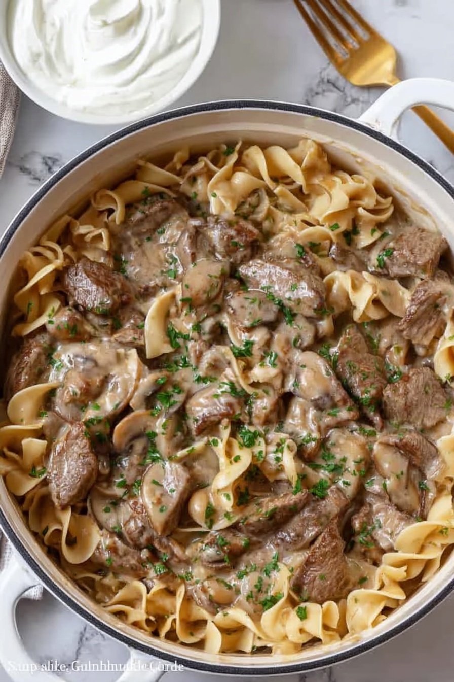 A white pot filled with a thick brown sauce that has visible pieces of orange carrots and sliced mushrooms within it. On top of the sauce, there is a pile of dark brown cooked strips of meat, arranged loosely in the center. The pot is placed on a white marbled surface. Nearby are two silver salt and pepper shakers and a white cloth with light gray stripes partially visible on the left side. photo taken with an iphone --ar 2:3 --v 7 - Creamy Beef Stroganoff, beef stroganoff recipe, easy beef stroganoff, hearty beef dinner, comfort food recipes