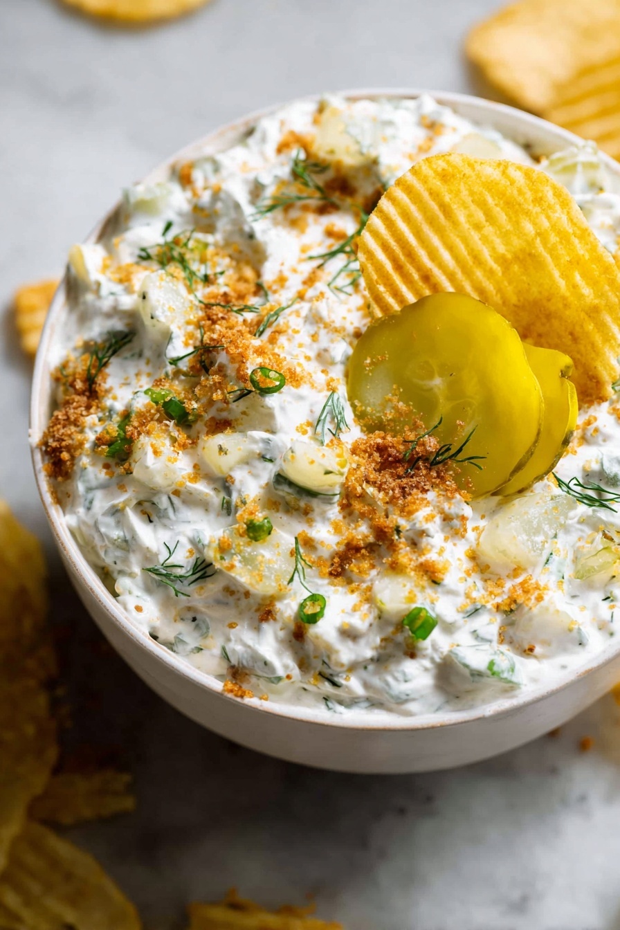 A white bowl filled with a creamy white dip mixed with small green herb pieces and light yellow chunks, topped with bright yellow pickle slices near the center. The dip is sprinkled with light brown crumbs and small green onion pieces, with a few small green herb sprigs scattered on top. A ridged golden potato chip is dipped into the creamy mixture near the edge of the bowl. The bowl sits on a white marbled surface with some crumbs and chips around it. photo taken with an iphone --ar 2:3 --v 7 - Ranch Fried Pickle Dip, fried pickle dip, quick appetizer dip, creamy dill dip, crispy panko pickle dip
