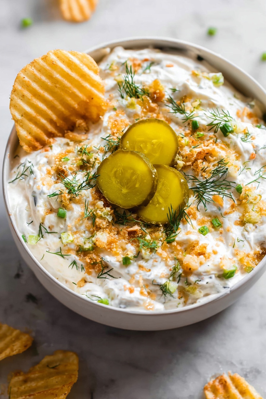 The image shows a grey bowl filled with a creamy white dip that is topped with several layers: small green chopped herbs, light brown crunchy bits, and three thick slices of pickles with a shiny, oily surface in the center. The dip looks thick and textured, with the crunchy bits scattered evenly on top. A woman’s hand is dipping a ridged, yellow chip into the creamy white mixture on one side. The bowl sits on a white marbled surface with some chips placed around it. photo taken with an iphone --ar 2:3 --v 7 - Ranch Fried Pickle Dip, fried pickle dip, quick appetizer dip, creamy dill dip, crispy panko pickle dip