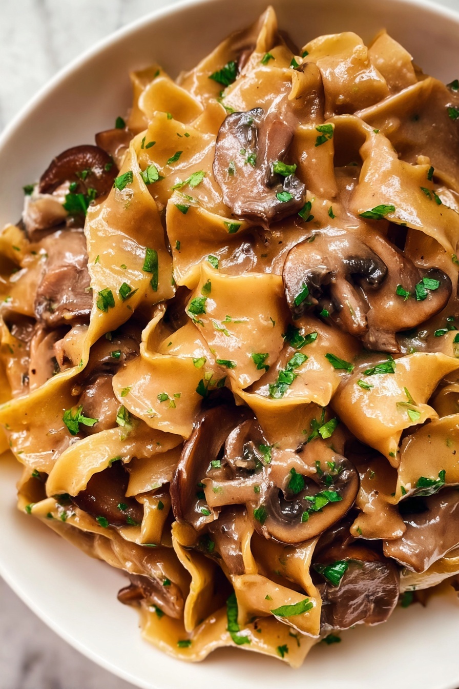 The image shows a close-up of a creamy pasta dish with wide, flat egg noodles mixed with thick slices of brown mushrooms. The noodles are coated in a smooth, light brown sauce that looks rich and glossy. Small pieces of chopped green herbs are sprinkled over the top, adding a fresh touch to the dish. The textures of the soft noodles and tender mushrooms blend together, showing a warm and comforting meal. The dish is served in a white bowl placed on a white marbled surface. photo taken with an iphone --ar 2:3 --v 7 - Vegetarian Mushroom Stroganoff, mushroom stroganoff recipe, vegan mushroom stroganoff, creamy mushroom pasta, meat-free comfort food