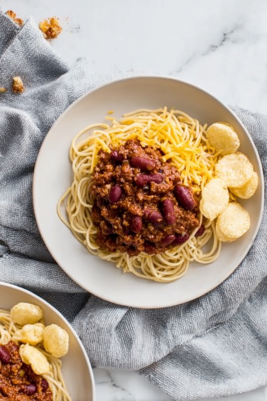 The image shows a white plate with three main layers. The bottom layer is a pile of light brown spaghetti noodles arranged in a loose small mound. On top of the noodles, there is a thick layer of dark reddish-brown chili with visible kidney beans and a chunky texture. To one side of the noodles and chili, there is a small group of round, pale beige crunchy chips neatly placed. The noodles and chips sit on a white marbled surface with a soft grey cloth draped around the plate. In the corner of the photo, part of another similar plate with the same food is visible. Photo taken with an iphone --ar 2:3 --v 7 - Cincinnati Chili Spaghetti, Cincinnati Chili, Cincinnati Chili Spaghetti recipe, Midwest chili dish, Cincinnati-inspired pasta