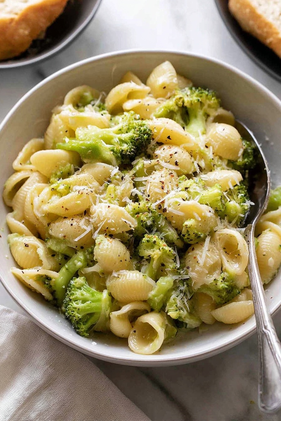 The image shows a white bowl filled with small shell pasta mixed with bright green broccoli pieces. The pasta looks soft and is coated with a light creamy sauce that makes the dish look smooth and shiny. There is a sprinkle of finely grated cheese on top and tiny specks of black pepper scattered over the dish. A silver spoon is placed partially inside the bowl on the right side. The bowl is set on a white marbled surface with a light cloth nearby and a blurred part of another dish with bread in the background. Photo taken with an iphone --ar 2:3 --v 7 - Quick Broccoli Pasta with Lemon and Cheese, healthy broccoli pasta, easy lemon pasta recipe, quick weeknight dinner ideas, cheesy garlic pasta