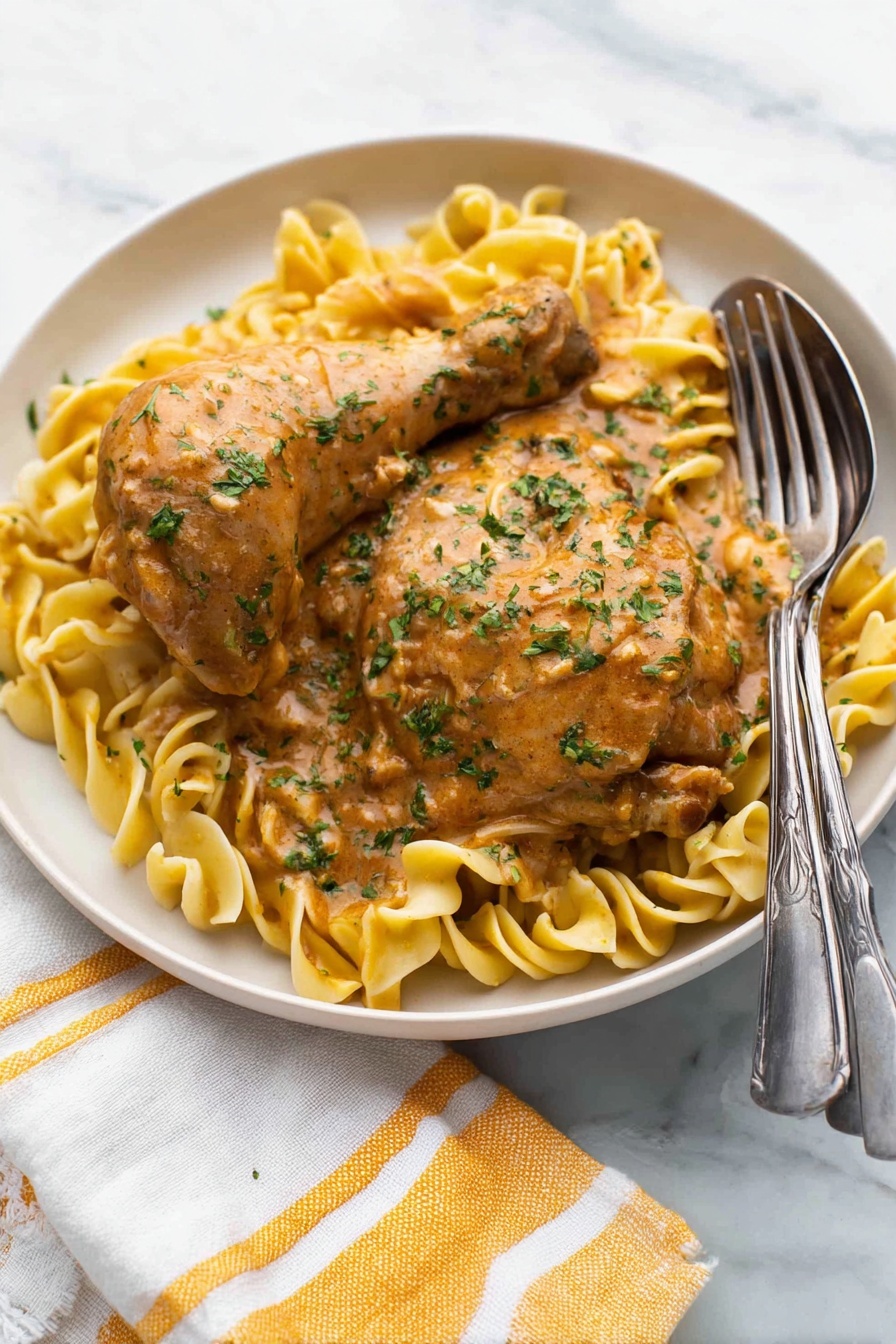 A white round plate holds two pieces of cooked chicken, one drumstick and one thigh, both covered in a thick light brown sauce with green herbs sprinkled on top. Underneath, there is a bed of light yellow egg noodles twisted softly, also coated lightly with the same sauce. A silver fork and spoon rest on the right edge of the plate. The plate sits on a white marbled surface with a white and yellow striped cloth napkin partially visible at the bottom left corner. Photo taken with an iphone --ar 2:3 --v 7 - Easy Chicken Paprikash, Hungarian Chicken Paprikash, Creamy Chicken Paprikash, Authentic Chicken Paprikash, Quick Chicken Paprikash