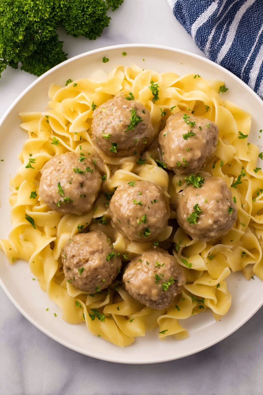 A white plate holds a dish with two main layers: six smooth, round beige meatballs evenly spread around the plate and a bed of soft, folded yellow egg noodles underneath and among the meatballs. The noodles have a slightly glossy texture and appear coated in light gravy. Both meatballs and noodles are lightly sprinkled with finely chopped green parsley, adding a fresh touch of color. The plate is set on a white marbled surface with a small bunch of parsley in the top left corner and a blue-striped towel in the bottom left corner. Photo taken with an iphone --ar 2:3 --v 7 - Creamy Swedish Meatball Noodle Skillet, Swedish meatball skillet, creamy meatball dish, one-pan Swedish meatballs, comfort food recipes
