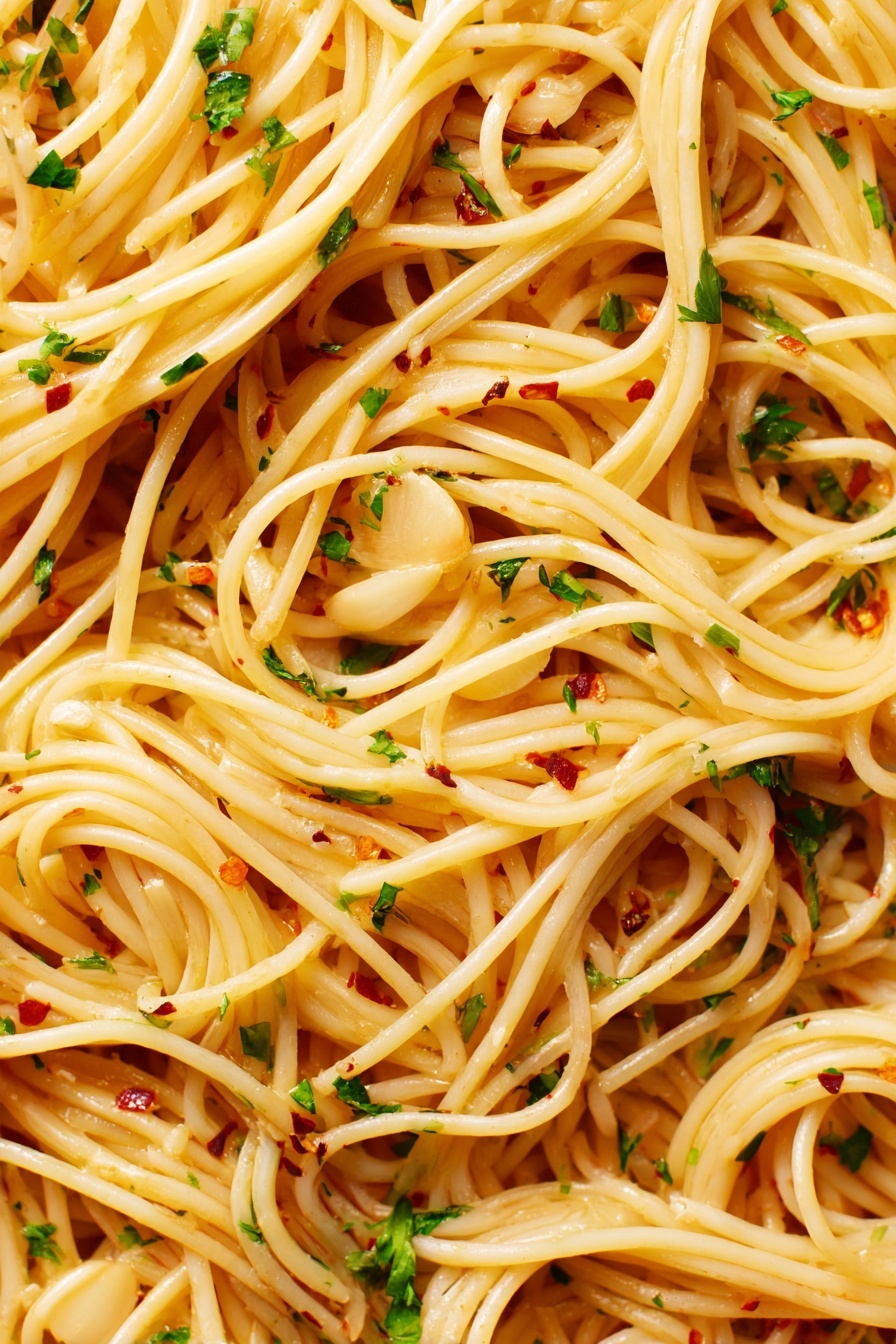The image shows a close-up view of spaghetti pasta with thin, long noodles that are light golden in color. The noodles are mixed evenly with thin slices of light tan garlic, small bits of green parsley, and tiny red chili flakes scattered throughout. The spaghetti appears glossy with a slightly oily texture, making the noodles look smooth and shiny. The background is a white marbled texture. photo taken with an iphone --ar 2:3 --v 7 - Garlic Spaghetti Aglio e Olio, easy garlic pasta, quick spaghetti recipes, simple Italian pasta, flavorful garlic spaghetti