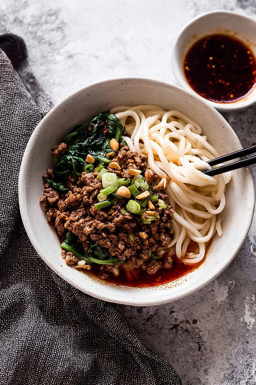 A white bowl filled with three main layers: at the bottom, thick, white noodles partially covered with a red oily sauce; in the middle, a generous layer of browned minced meat topped with chopped green onions and crushed peanuts; and on the top, fresh green leafy vegetables arranged on one side. A pair of wooden chopsticks held by a woman's hand is lifting some noodles, showing their texture and sauce coating. The bowl is placed on a white marbled surface with scattered green onion pieces and crushed peanuts nearby. photo taken with an iphone --ar 2:3 --v 7 - Spicy Dan Dan Noodles with Pork, easy Dan Dan noodles, homemade Dan Dan noodles, Sichuan pork noodle recipe, spicy noodle bowl