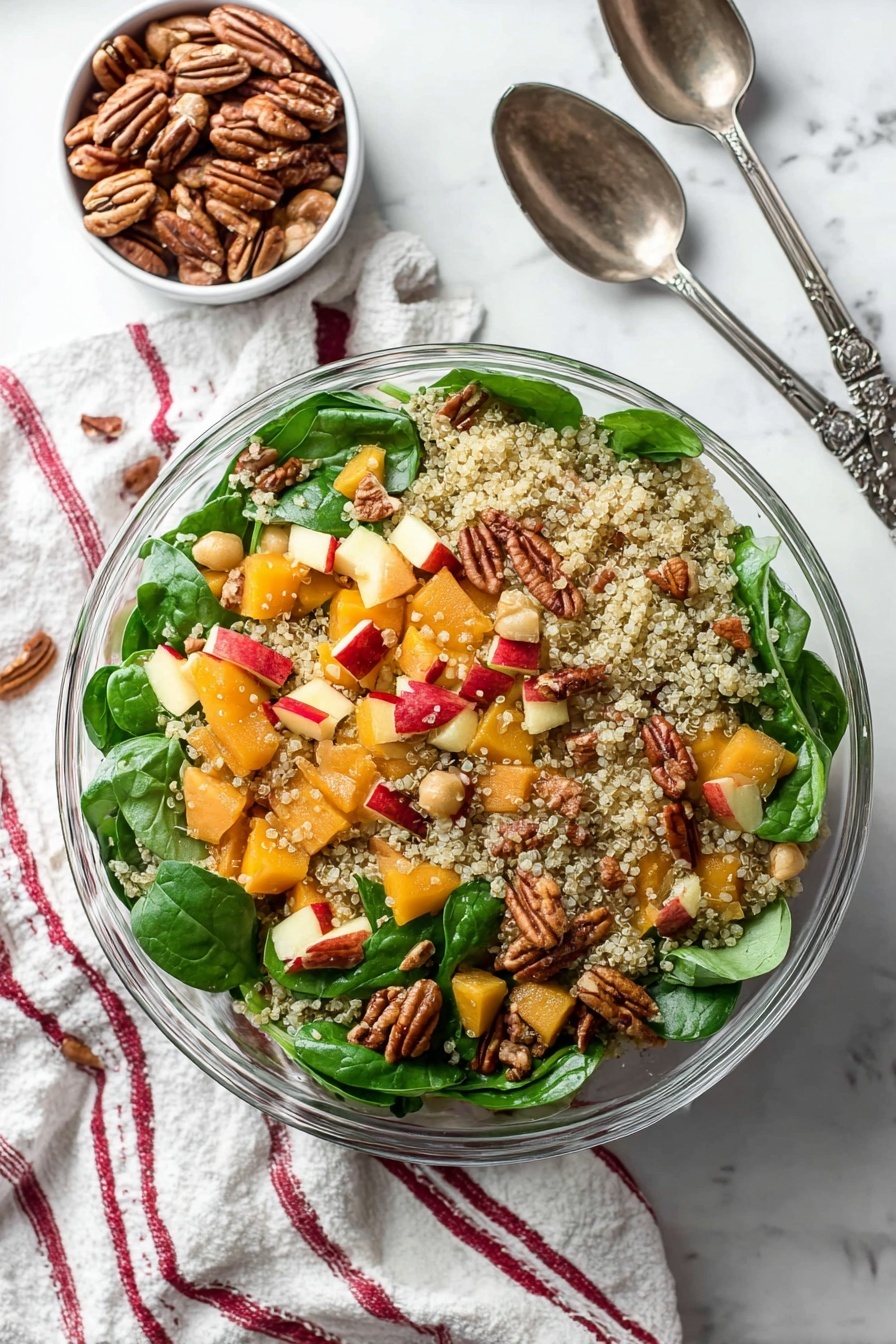 This image shows a clear glass bowl filled with a colorful quinoa salad. The base layer is light, fluffy quinoa with small, round grains scattered evenly throughout. Mixed inside are bright green fresh spinach leaves with a smooth texture, orange chunks of butternut squash, and small pale beans adding softness. Red-skinned apple pieces give a splash of red color, and whole pecans add a textured, brown, nutty layer randomly placed on top. The bowl is on a white marbled surface with a soft striped cloth nearby, and a woman's hand holds a dark metal salad spoon at the edge. photo taken with an iphone --ar 2:3 --v 7 - Roasted Butternut Squash Quinoa Salad, fall quinoa salad, healthy roasted veggie salad, vegan quinoa salad, cozy fall salad