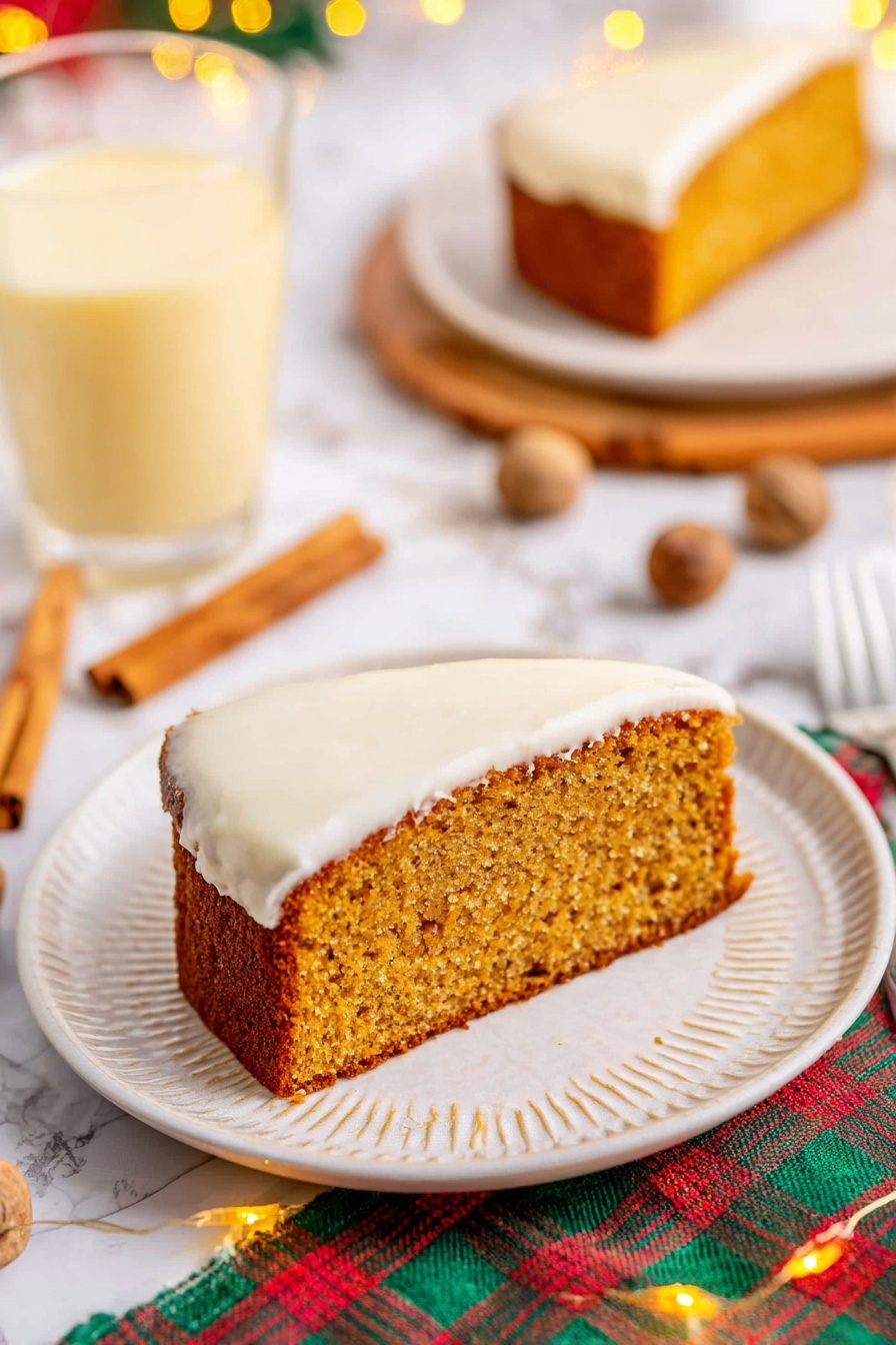 The image shows a single slice of moist, golden-brown cake with a thick layer of smooth white frosting on the bottom side, sitting on a white plate with a subtle textured rim. The cake slice has a slightly rough texture on top, indicating it is soft inside. Around the plate, there are cinnamon sticks and whole nutmegs scattered on a white marbled surface, with a colorful plaid cloth partially visible underneath. In the background, there is a clear glass filled with a light yellow creamy drink, and a second plate holding another slice of the same cake. Small warm fairy lights add a cozy touch to the scene. Photo taken with an iphone --ar 2:3 --v 7 - Eggnog Bread with Spiced Glaze, festive eggnog bread, holiday bread recipes, spiced holiday bread, moist eggnog loaf